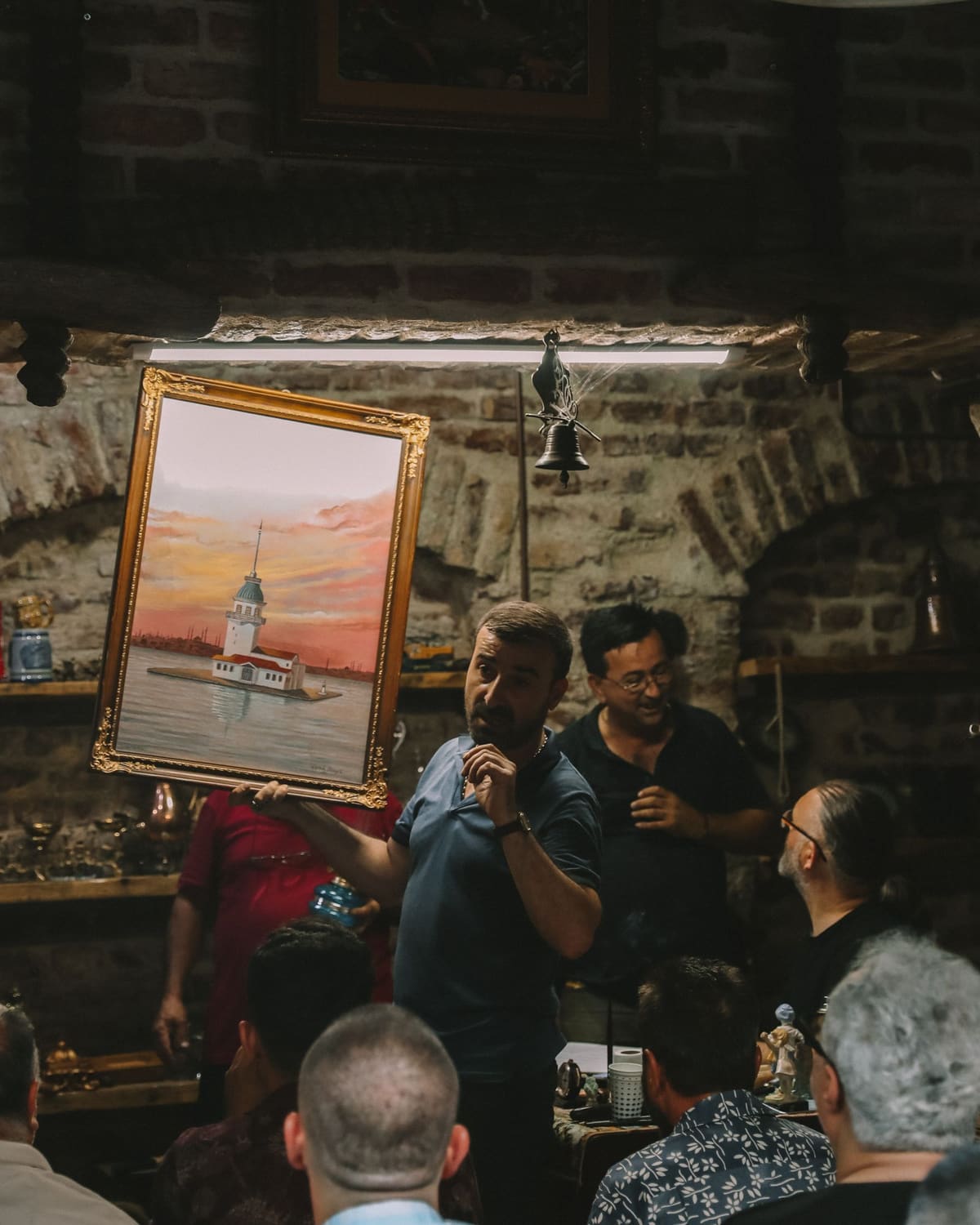 A man holding up a framed painting to an attentive crowd in a warm brick auction room, the auction bell overhead.