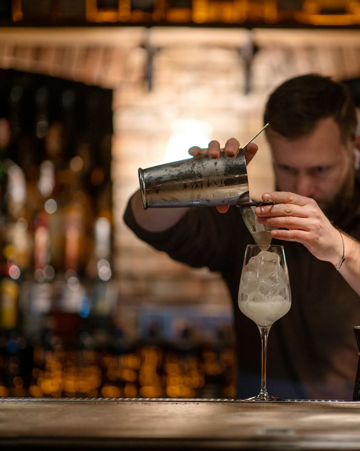 A bartender double-straining a cocktail into a glass in a warm, brick-walled bar