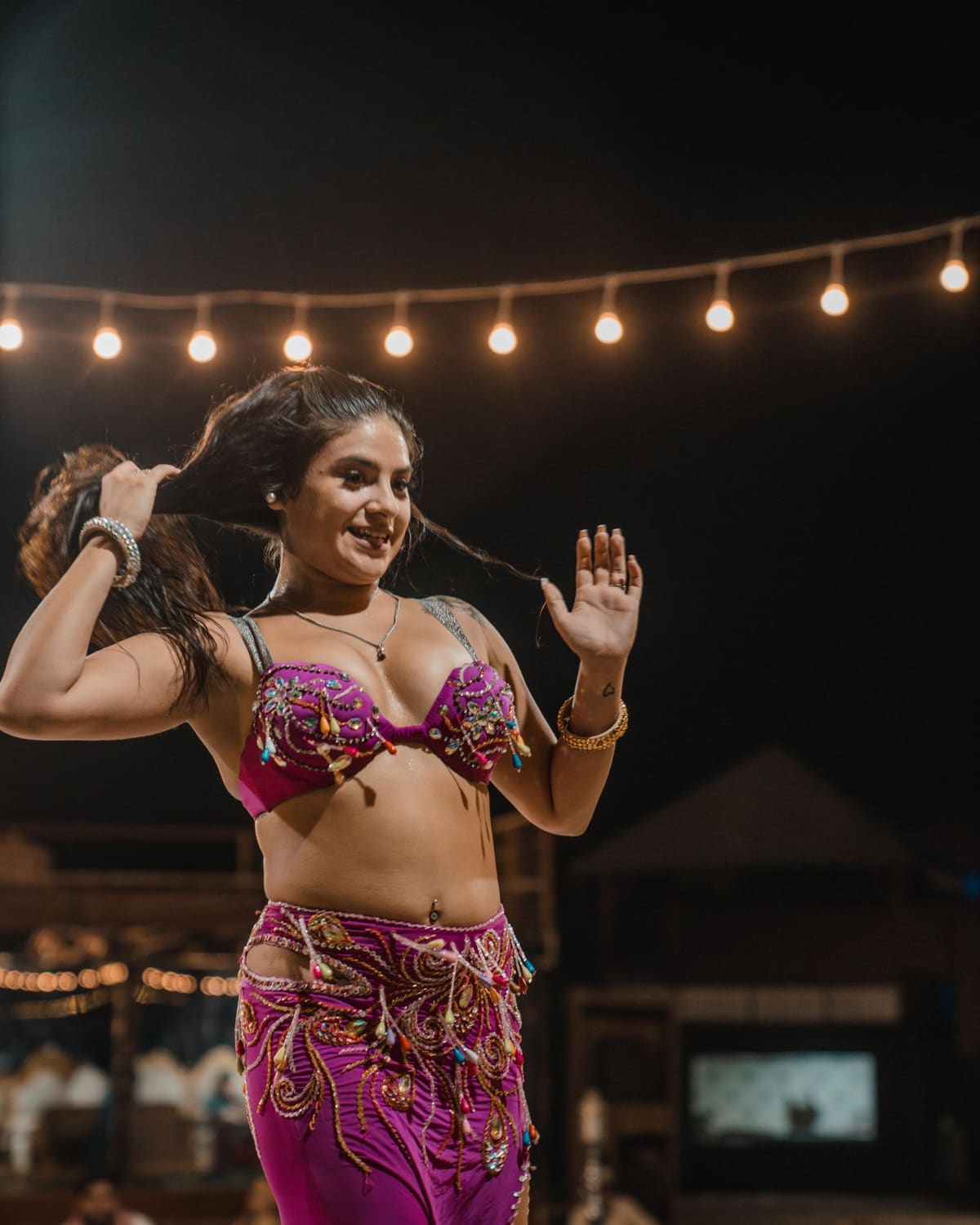 A belly dancer in a magenta costume smiles mid-move under string lights at night.