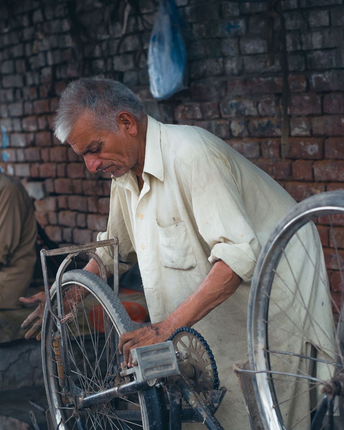 An older man in a light shirt bent over a bicycle wheel, repairing it against a brick wall in warm daylight.