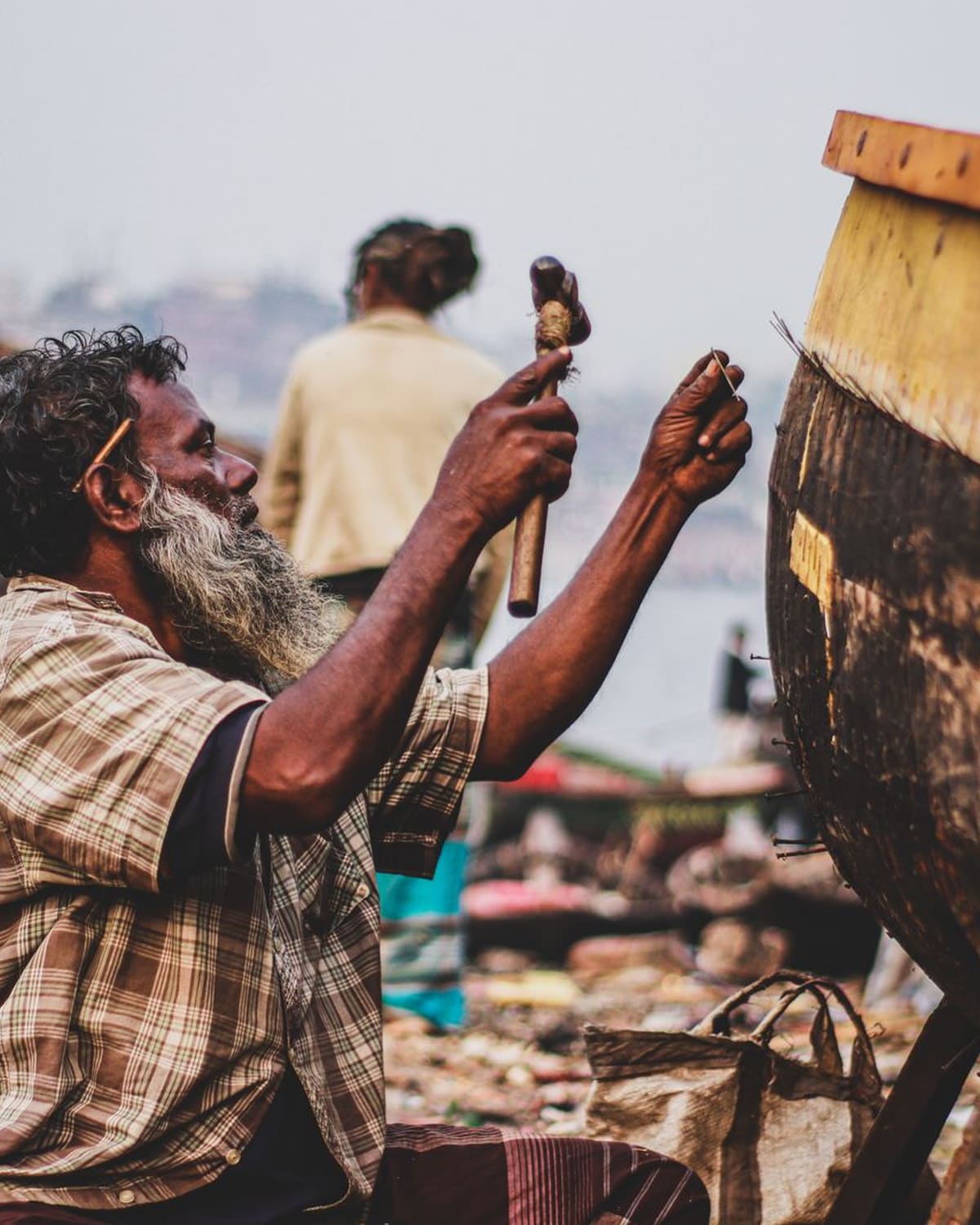 A bearded craftsman raising a mallet to a wooden boat hull at a riverside boatyard in soft daylight.