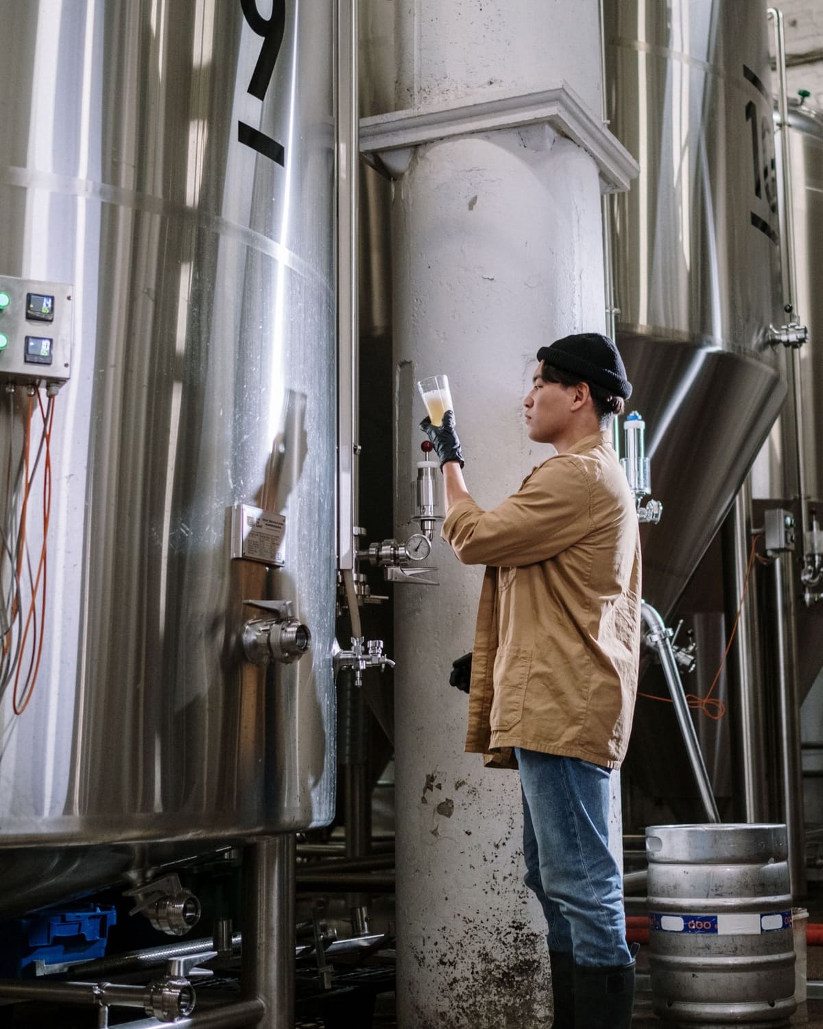 A bearded brewer in a plaid shirt and cap leaning over a large stainless steel brewing tank, adjusting the fittings on top.