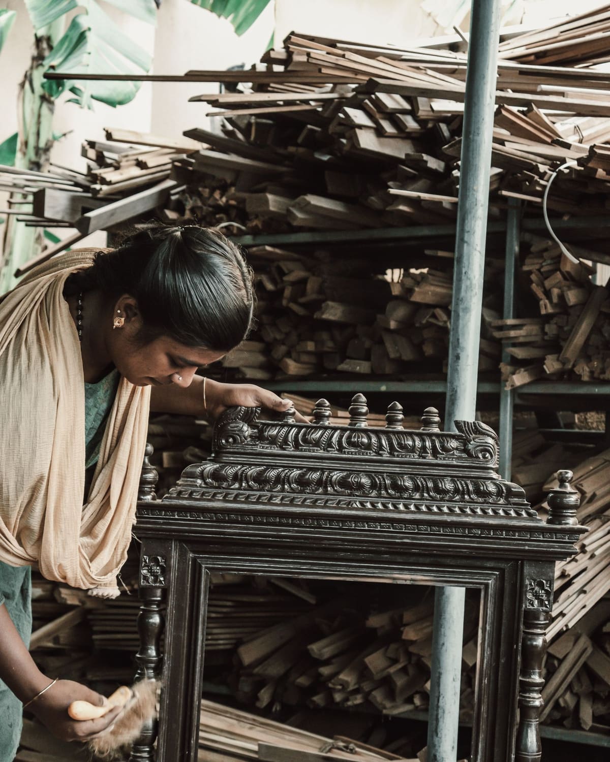 A woman bent over a carved dark wood cabinet in a workshop, brushing the frieze, with timber stacked to the roof behind her.