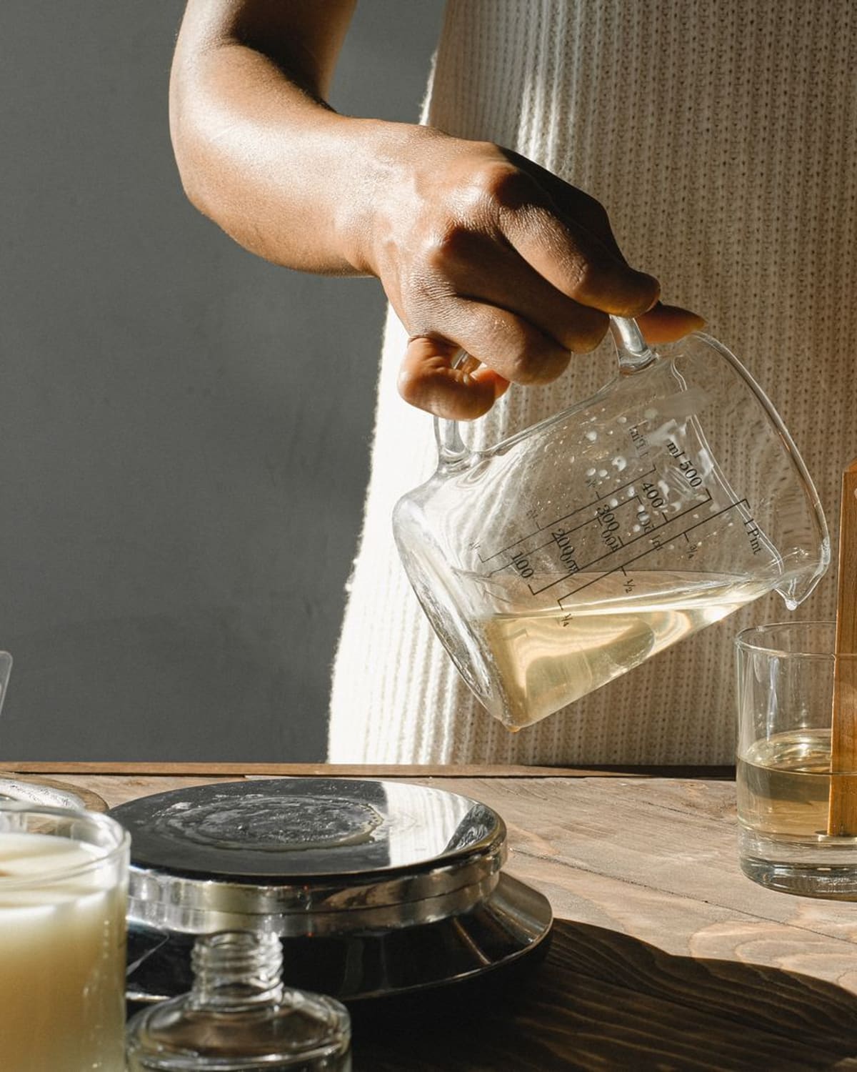 A woman's hands pouring melted candle wax from a glass jug into a glass holding a wooden wick, in bright window light on a wooden table.