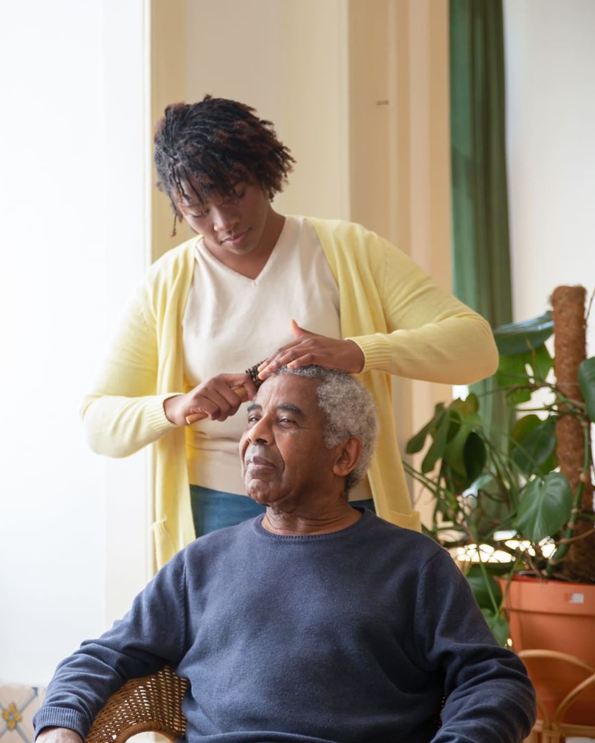 A caregiver gently combing an older man's hair by a sunlit window.