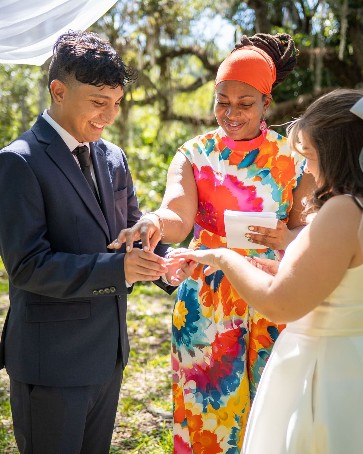 A celebrant in a bright floral dress guiding a couple through a ring exchange outdoors while everyone smiles.