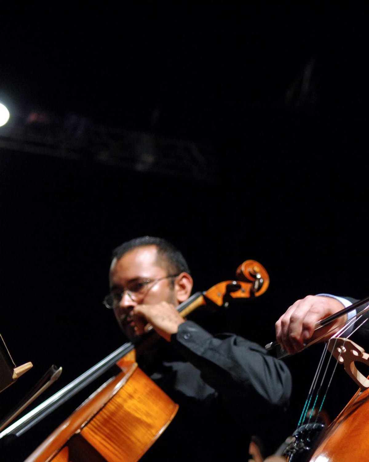 Two men in suits play cellos on a darkened stage, bows drawn across the strings, warm stage light on their faces and the wood of the instruments.