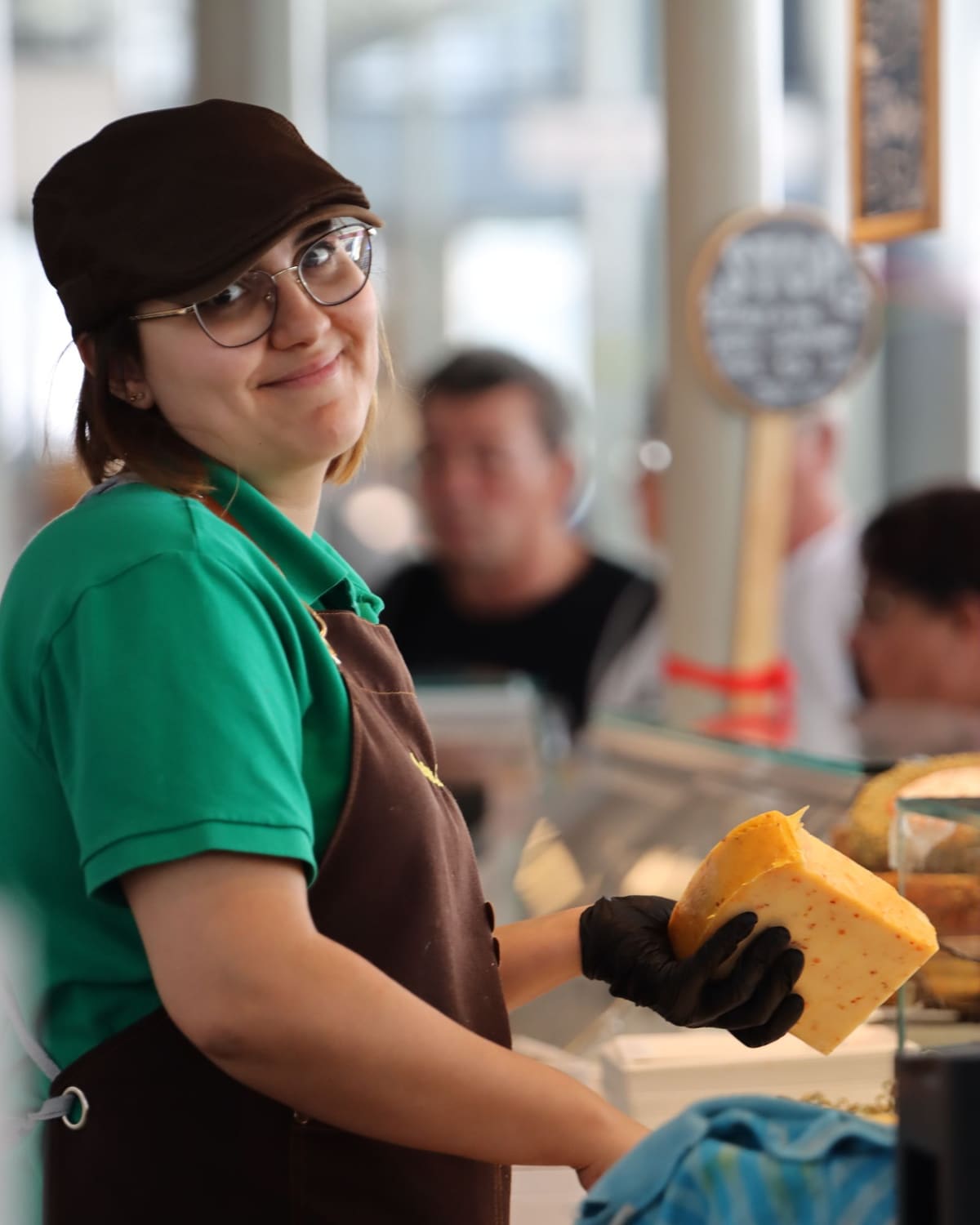 A smiling cheesemonger in an apron holding a wedge of cheese behind a market counter