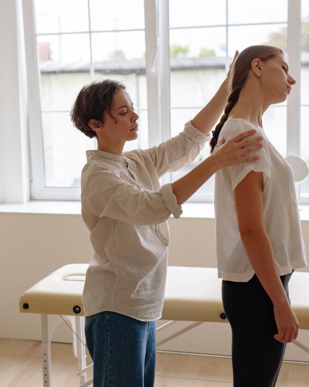 A practitioner with both hands on a standing clients neck and shoulder, checking their alignment in a bright room.