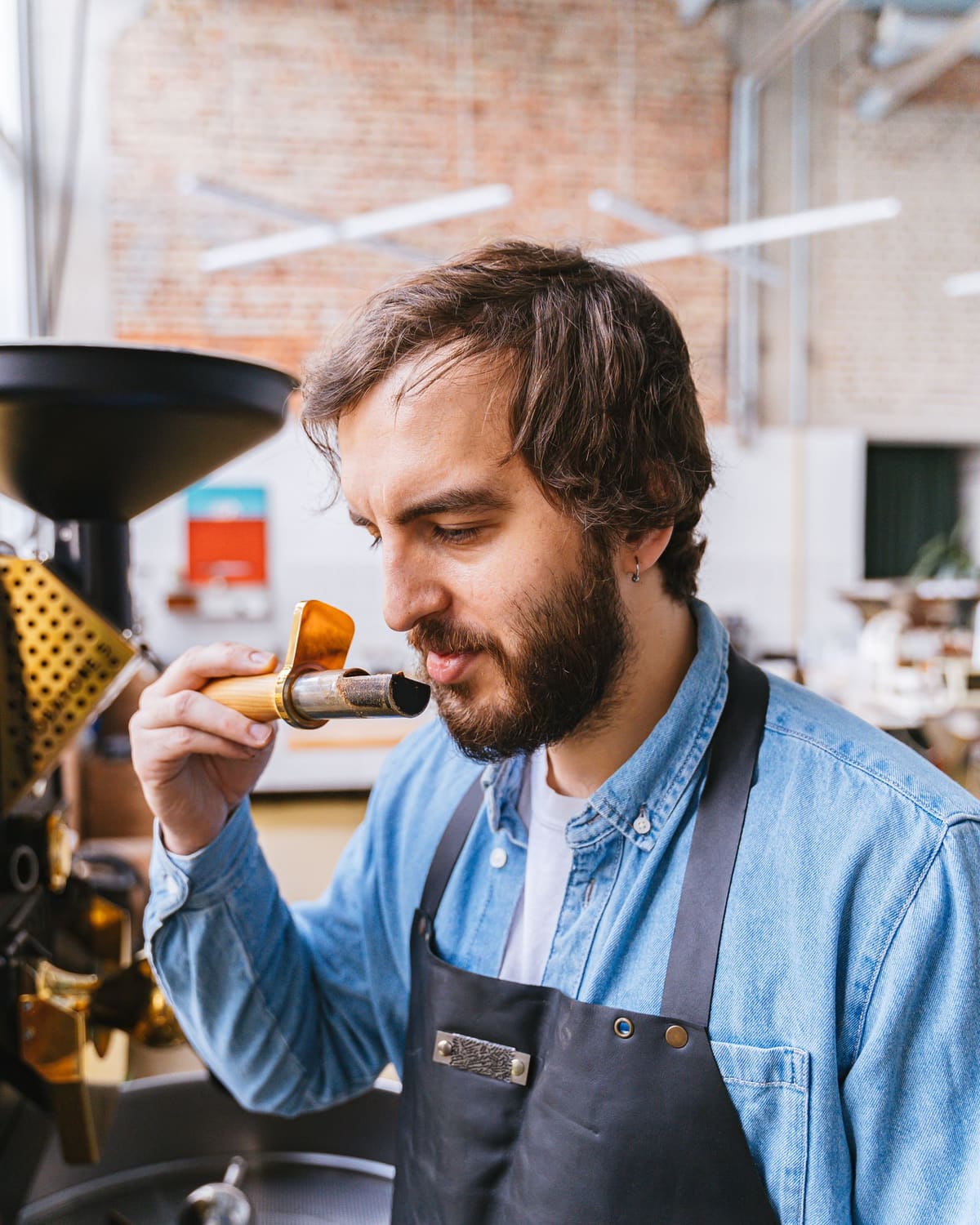 A bearded man in a denim shirt and apron smelling a sample scoop of freshly roasted beans beside a coffee roasting machine in a brick-walled roastery.