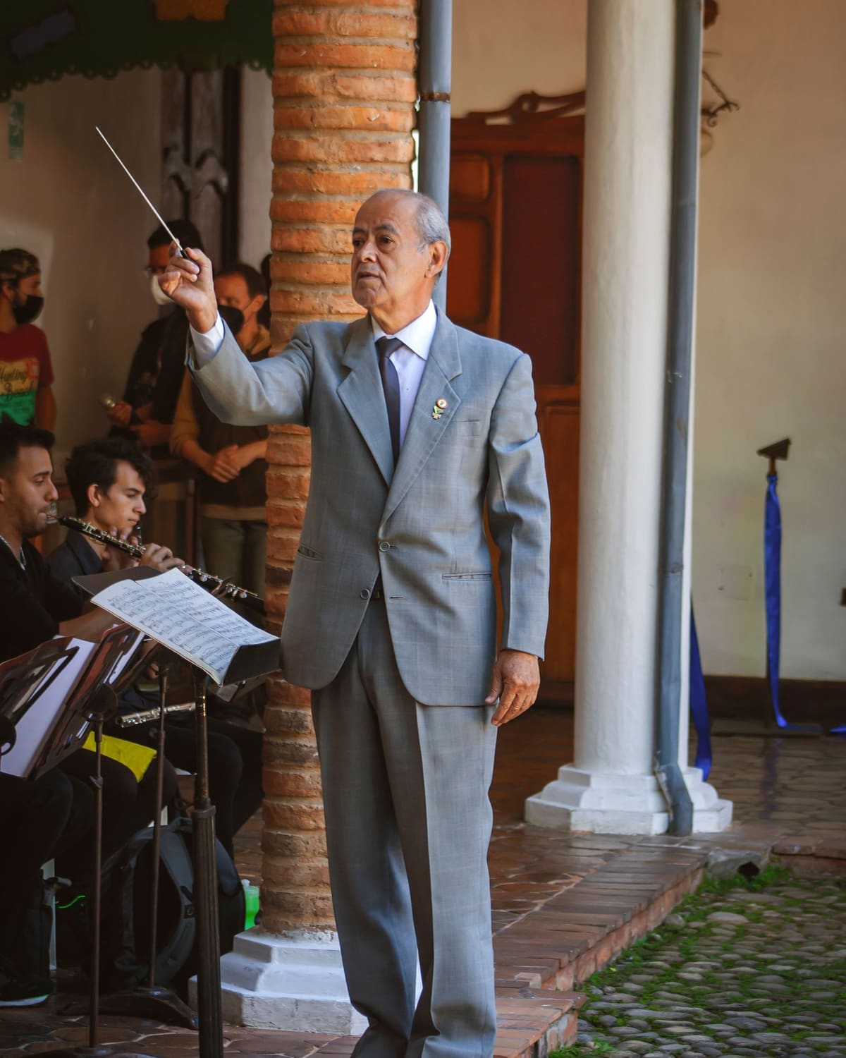 An older conductor in a light grey suit stands on a small podium with baton raised, leading young musicians who play wind instruments in a brick and stone courtyard.