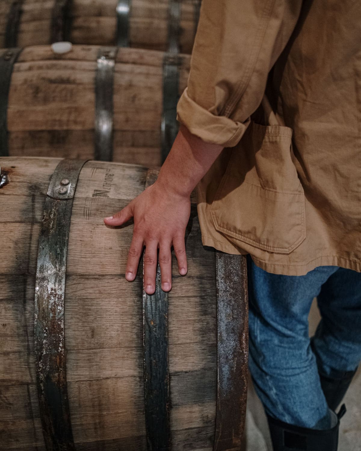 A cooper in a cap and work gloves bent over a row of oak whisky casks at a working cooperage.