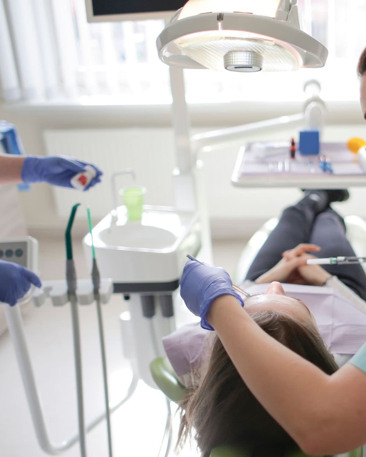 A dental hygienist in scrubs and mask leaning in to clean a reclined patient's teeth with fine tools