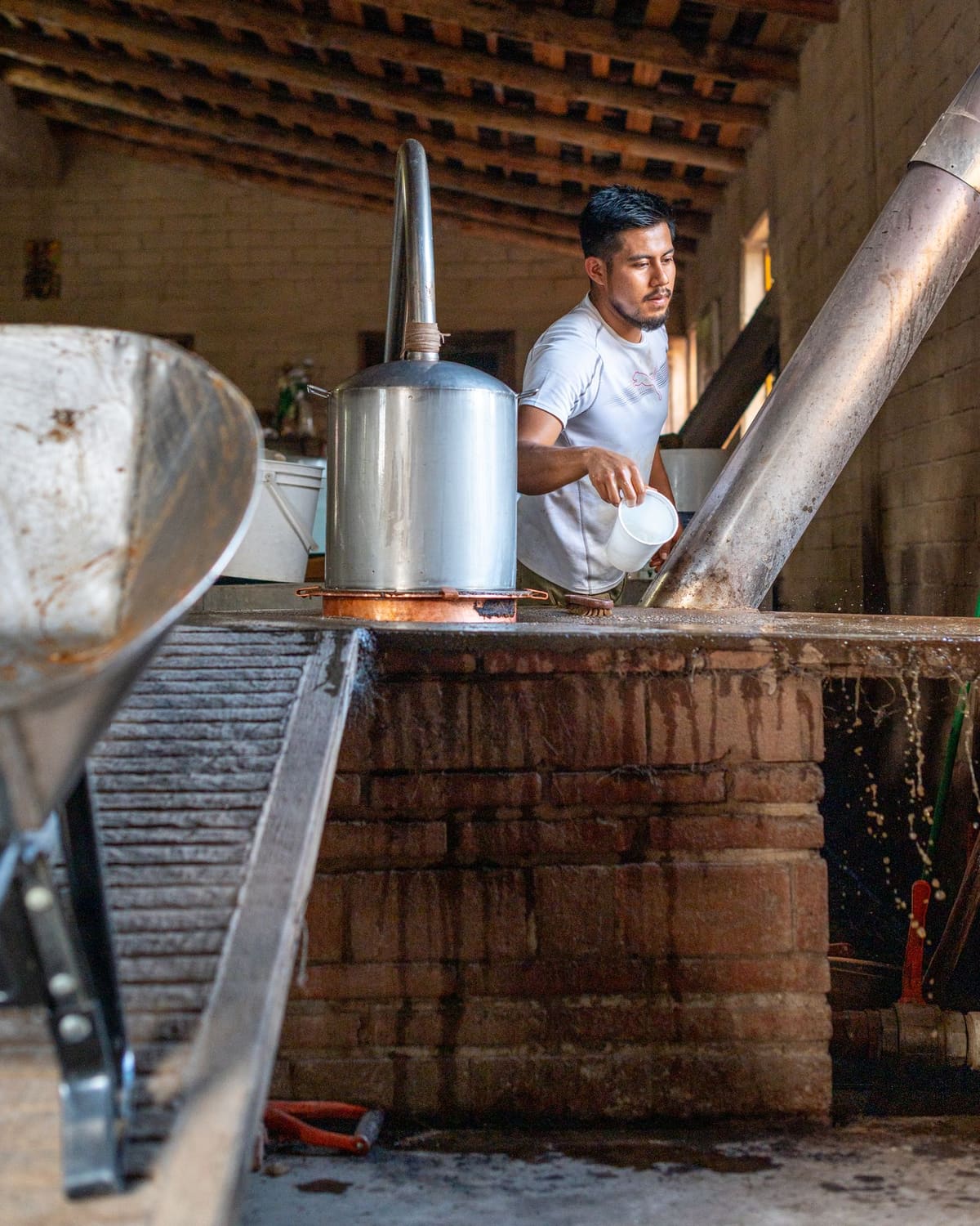 A young man in a white shirt tending a traditional agave still, pouring from a cup beside a metal pot still with a copper base and brick furnace, lit by a nearby window.