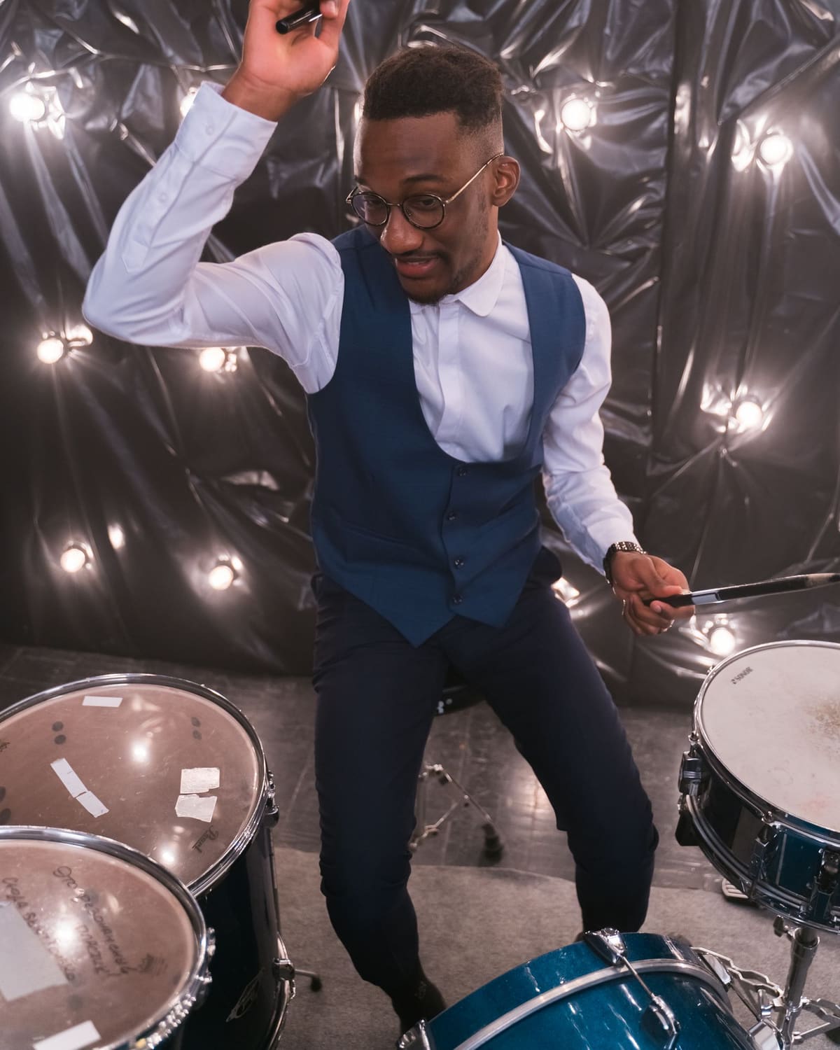 A drummer in a waistcoat swings a stick mid-beat at his kit under warm bulb lights.