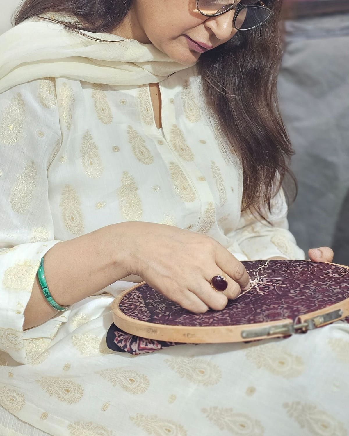 A woman in glasses and white clothing embroiders a maroon fabric stretched in a wooden hoop.