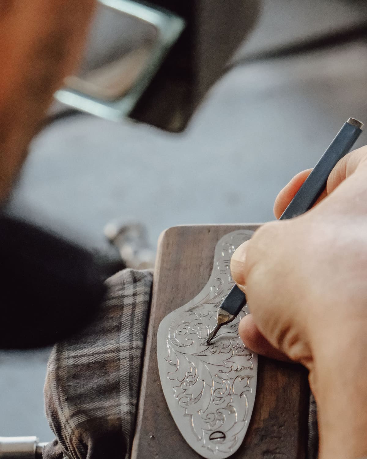 A hand pushes a graver to cut a fine scroll pattern into a silver piece held in a wooden block.