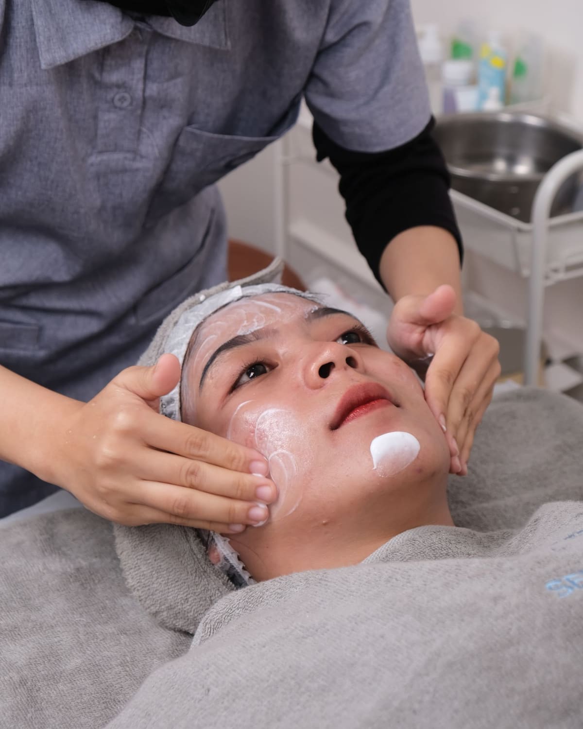 An esthetician gently applying cream to a reclining client's face with both hands during a facial.