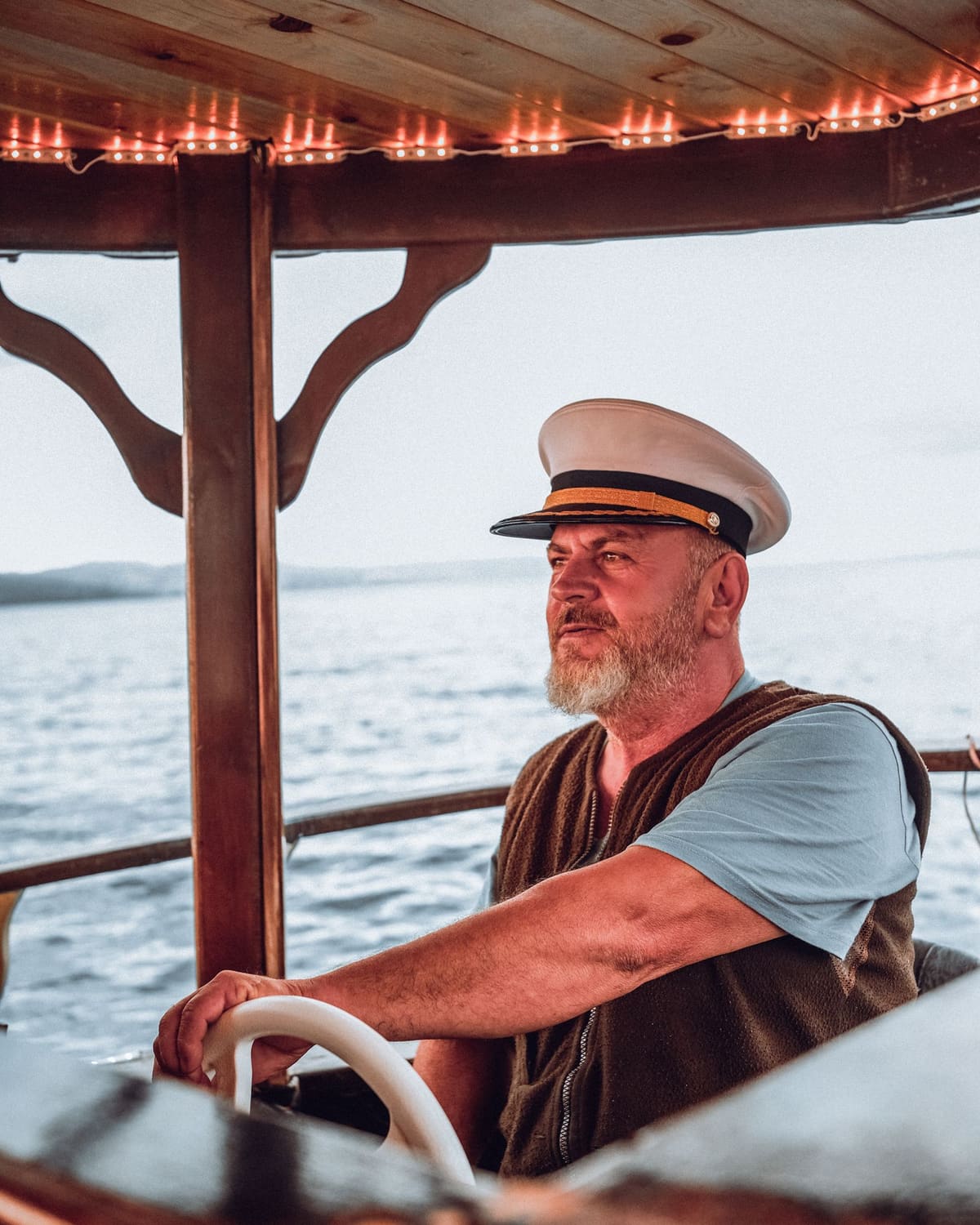 A ferry captain in his cap at the helm, water behind him