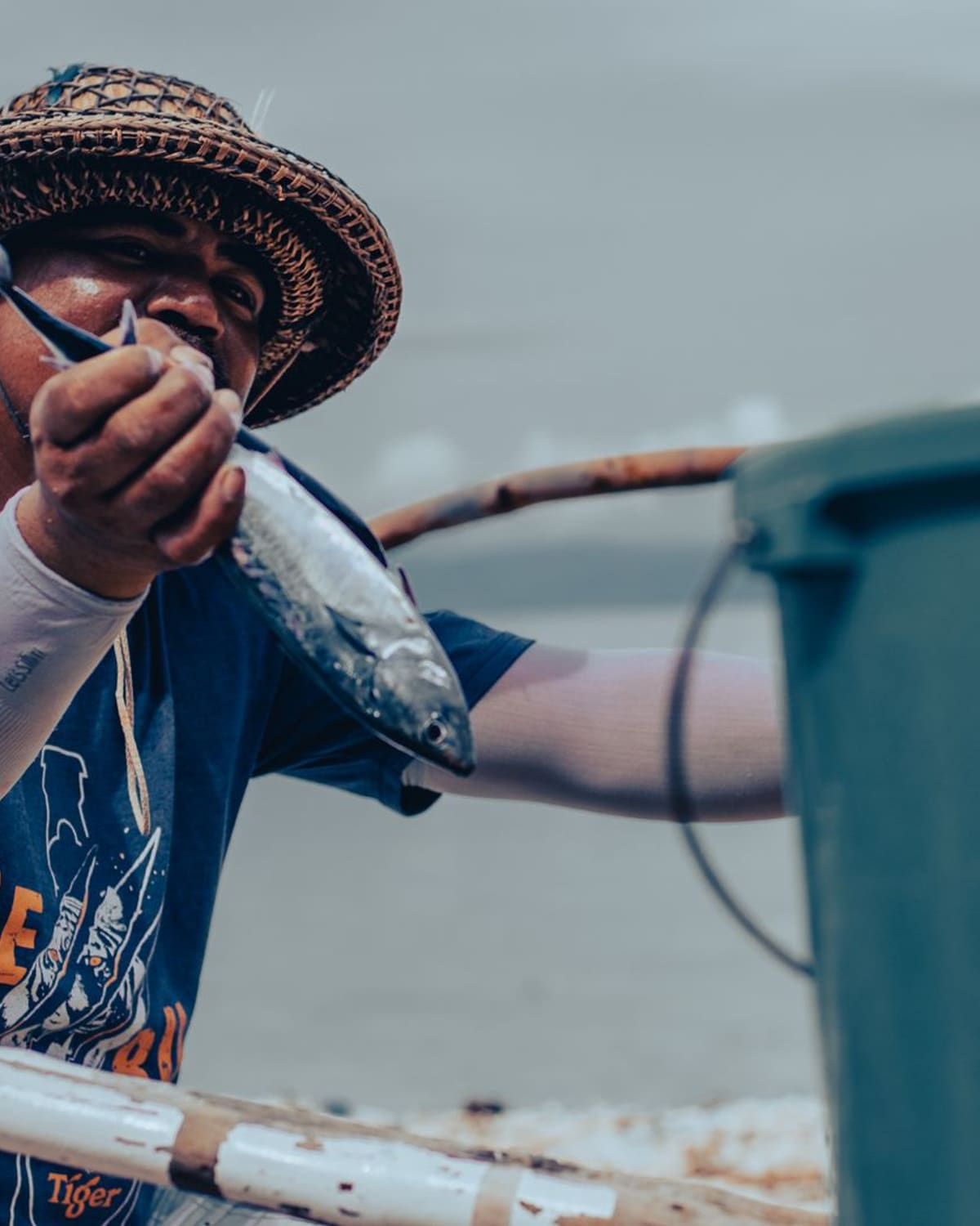 A fisherman in a straw hat on his boat holding up a freshly caught fish.