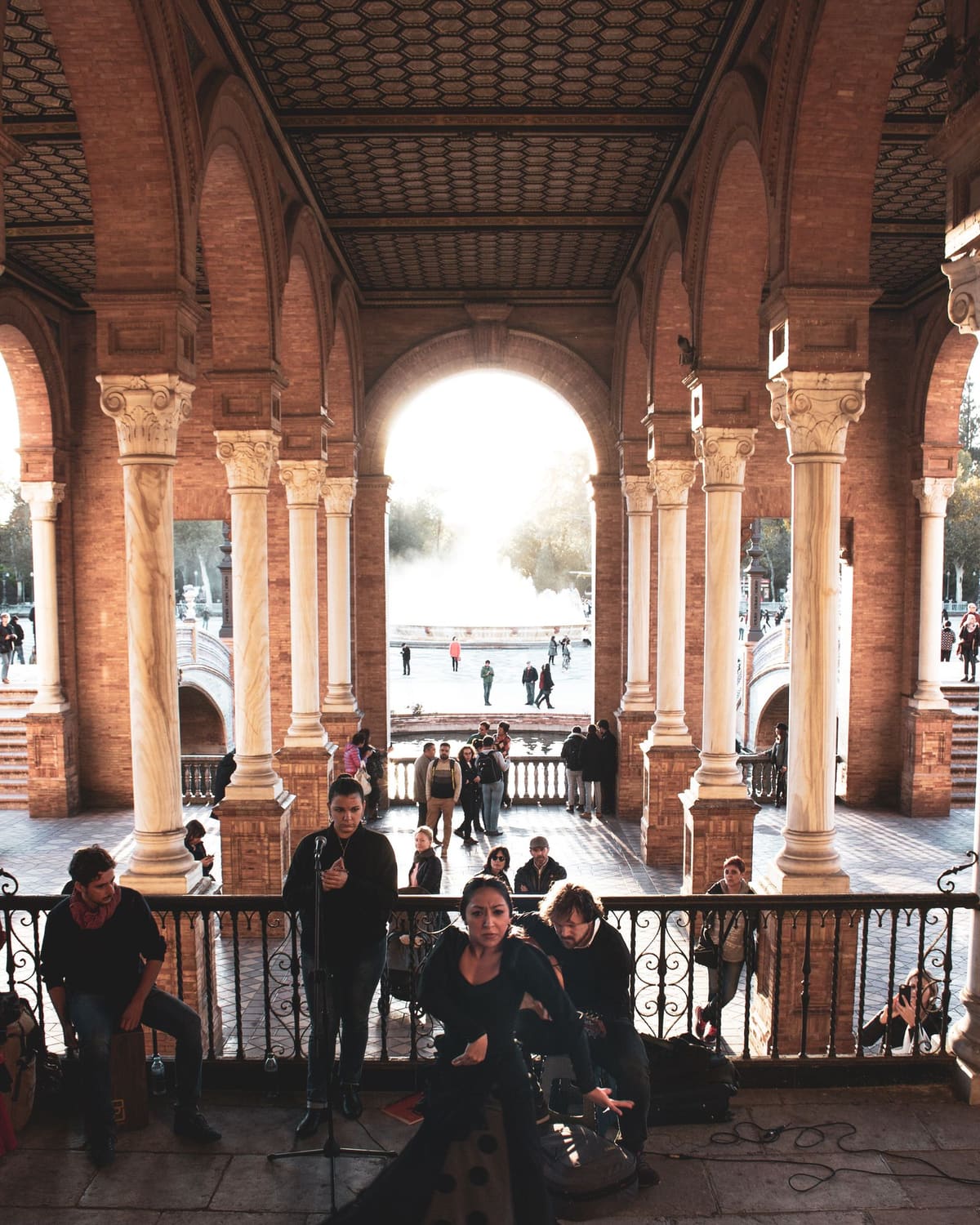 A woman in a long black flamenco dress dances on a stone terrace framed by grand arches, with a guitarist, a singer at a microphone, and onlookers behind her in golden light.