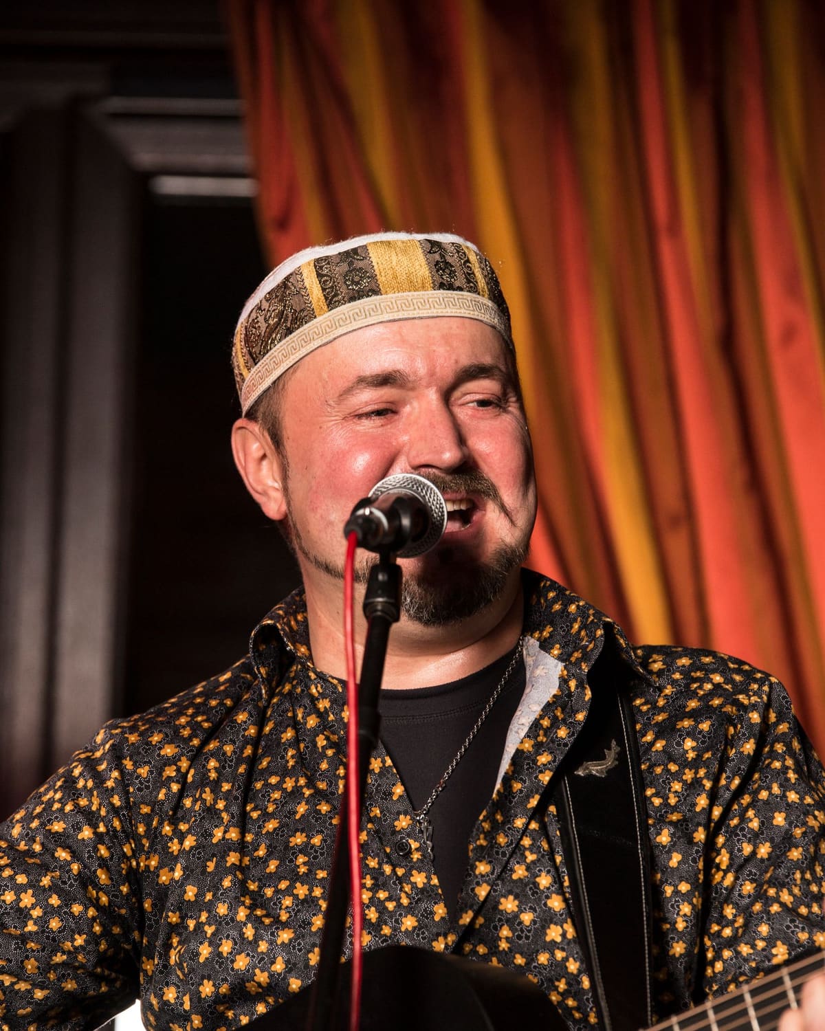 A folk singer in a cap sings into a microphone while playing an acoustic guitar under warm stage light.