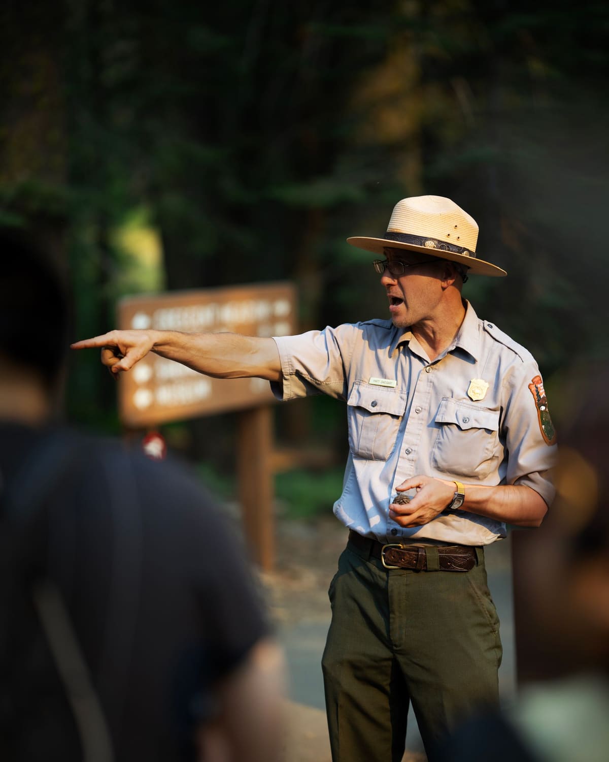 A uniformed ranger in a flat-brim hat pointing and speaking to visitors at the edge of a forest in golden light