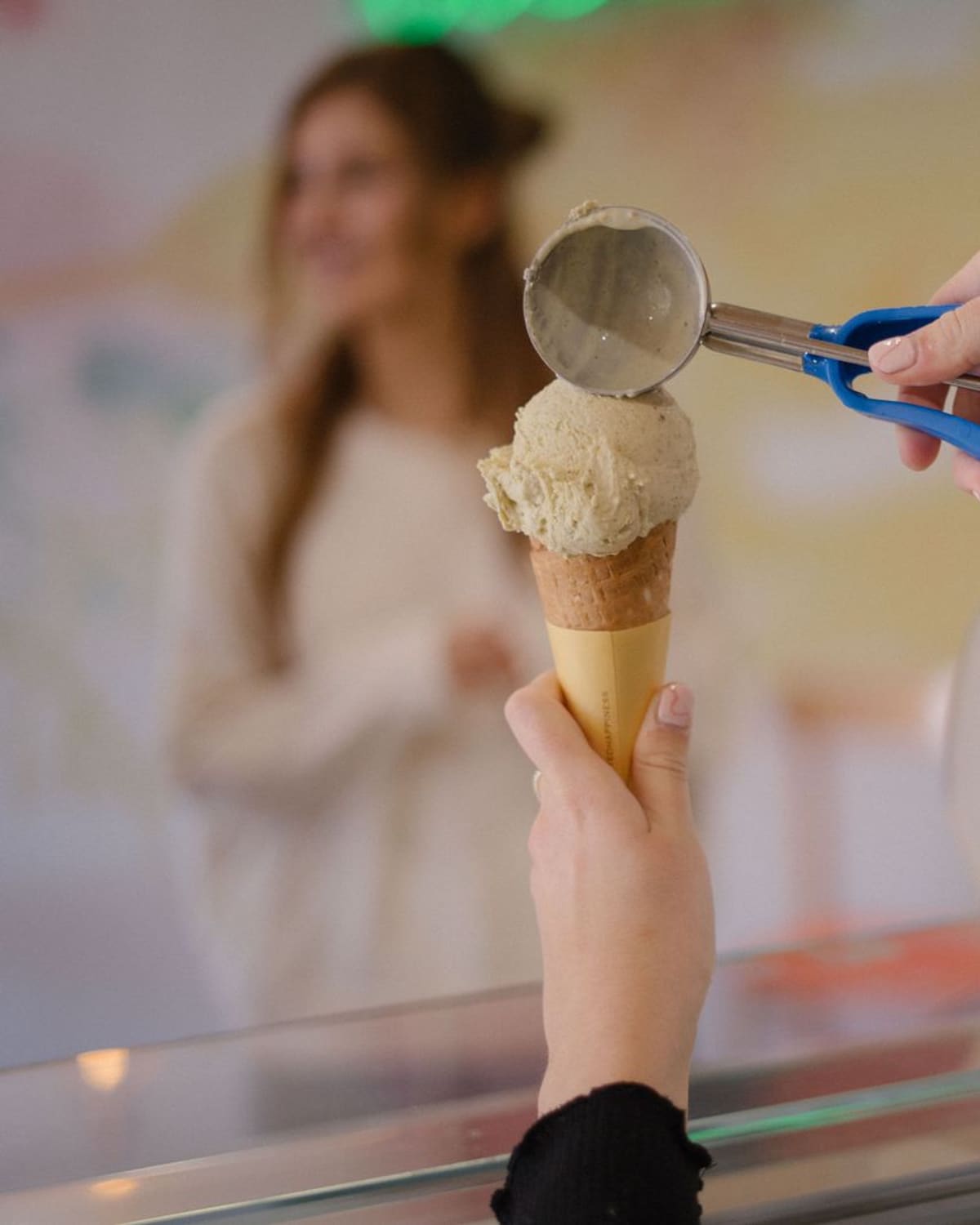 A server's hands scooping pale pistachio gelato onto a waffle cone at a gelato counter, with trays of colourful gelato in the case behind.