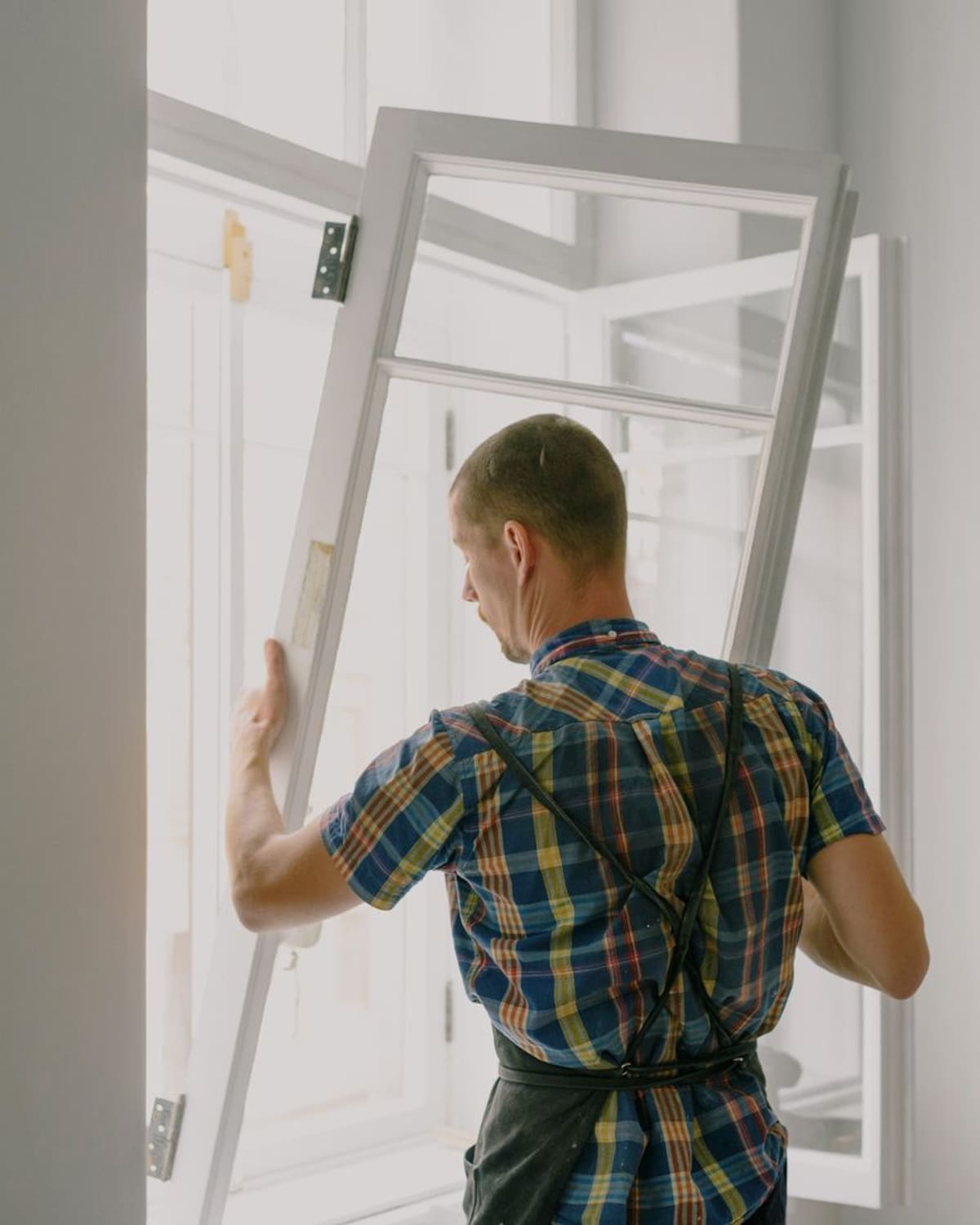 A man in a checked shirt seen from behind, lifting and fitting a white-framed glazed window in a bright room.