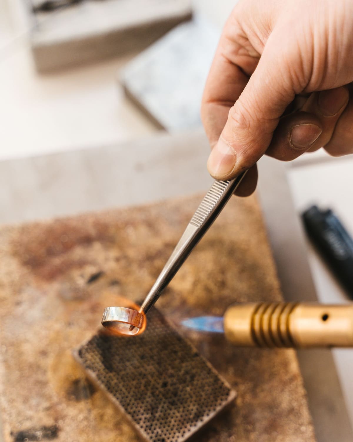 A jeweler's hand holds a ring in tweezers over a small flame on a soldering block.