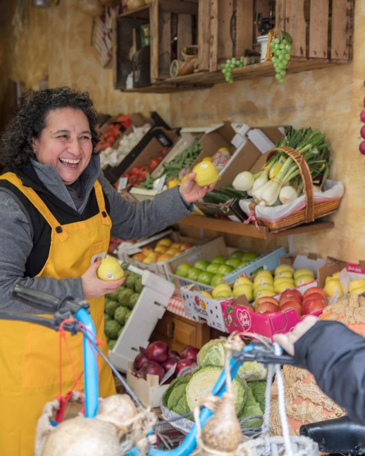 A smiling greengrocer in a yellow apron handing fruit across a colorful produce stall.