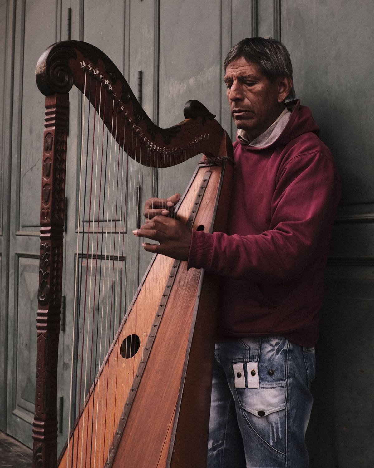 A man in a maroon hooded sweatshirt stands playing a tall carved wooden harp, both hands on the strings, in front of a pale grey panelled door.