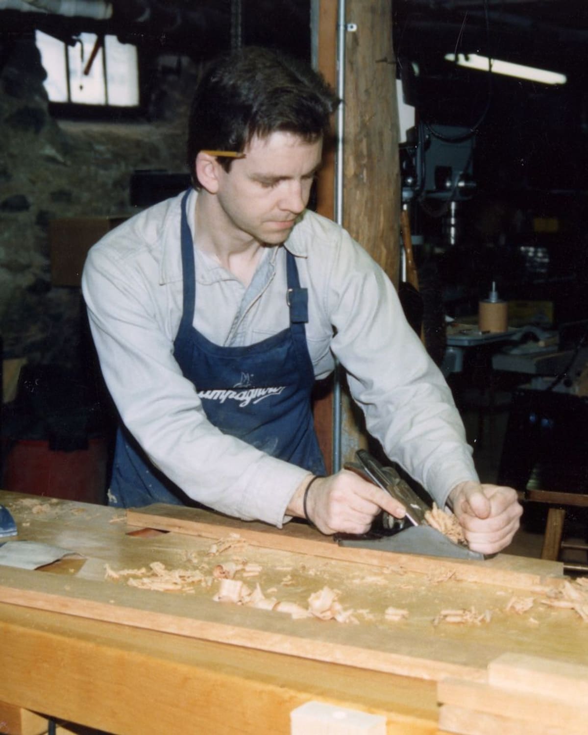 A craftsman at a workbench shaping timber by hand, shavings gathering around the plane.