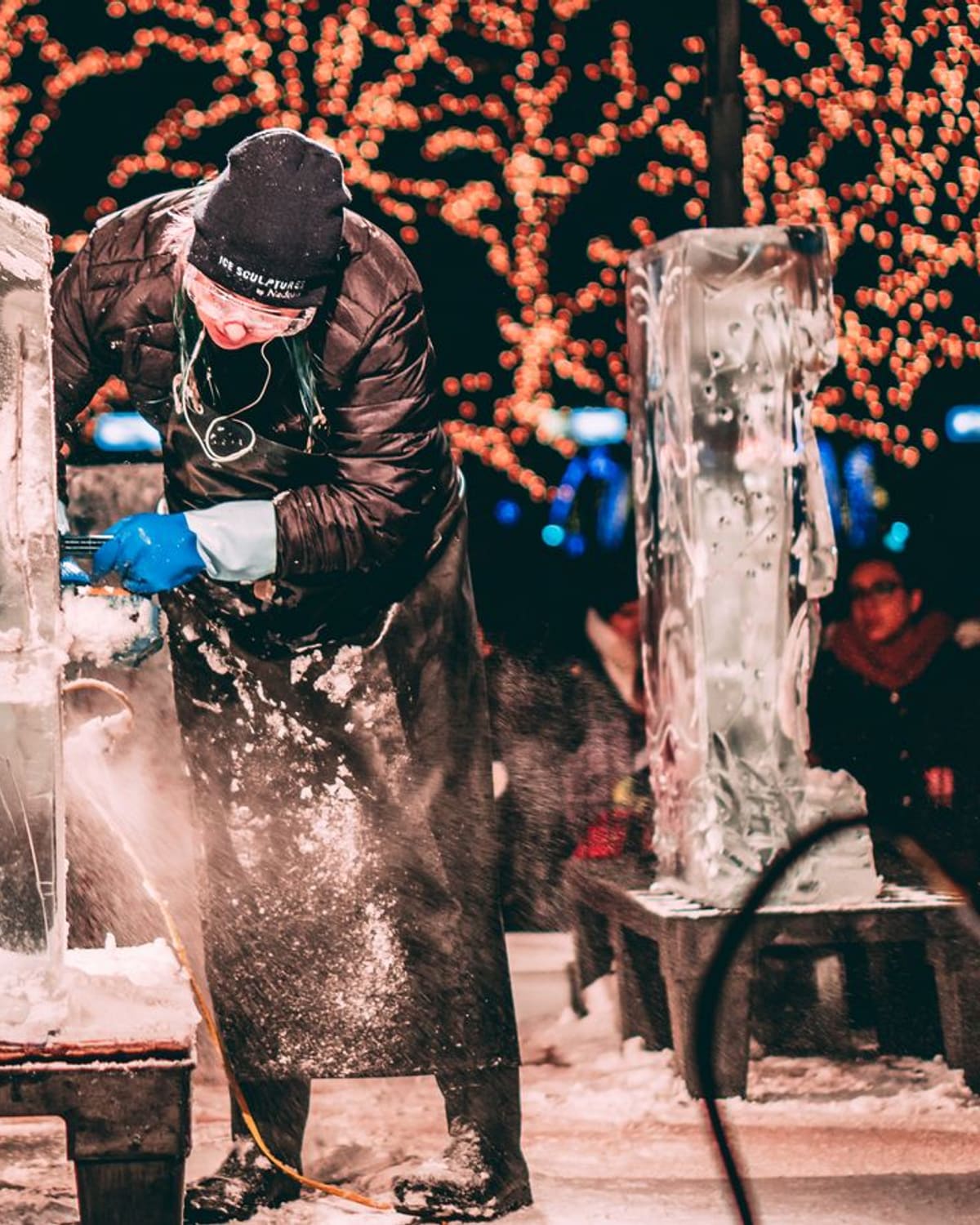 An ice sculptor bent over a block of ice carving it with a tool at a winter festival, warm lights behind