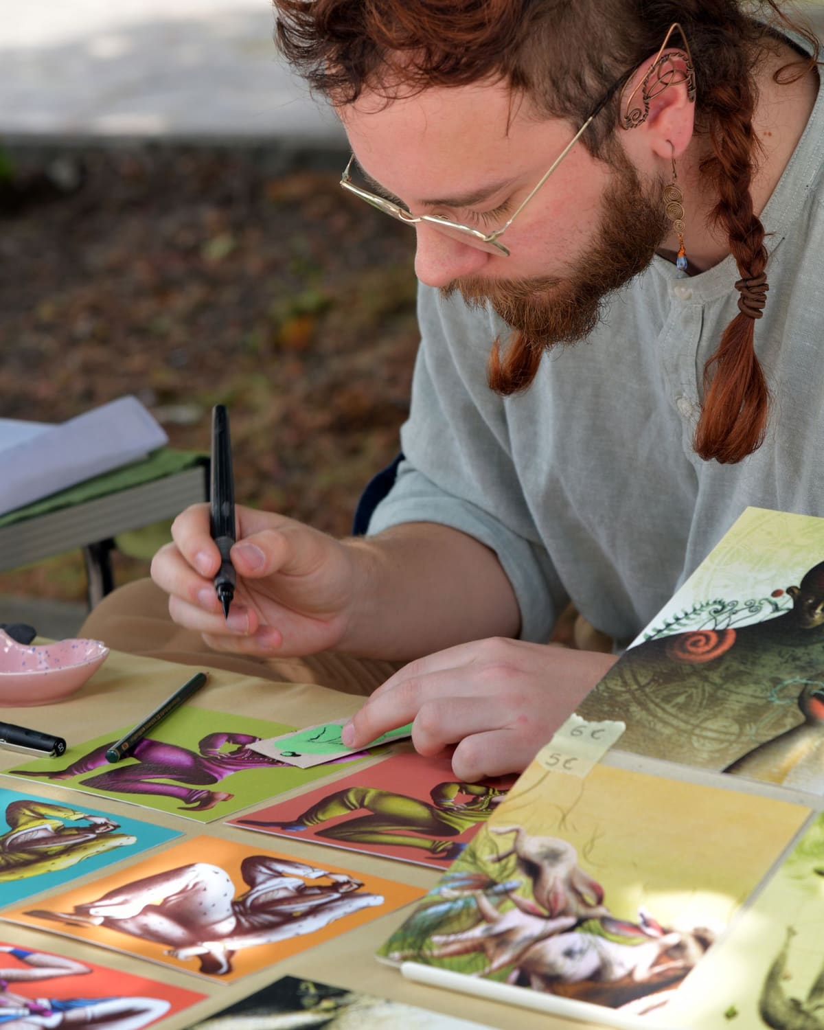 A young man with red hair draws in black ink at an outdoor table, with his finished illustrations spread out around him.