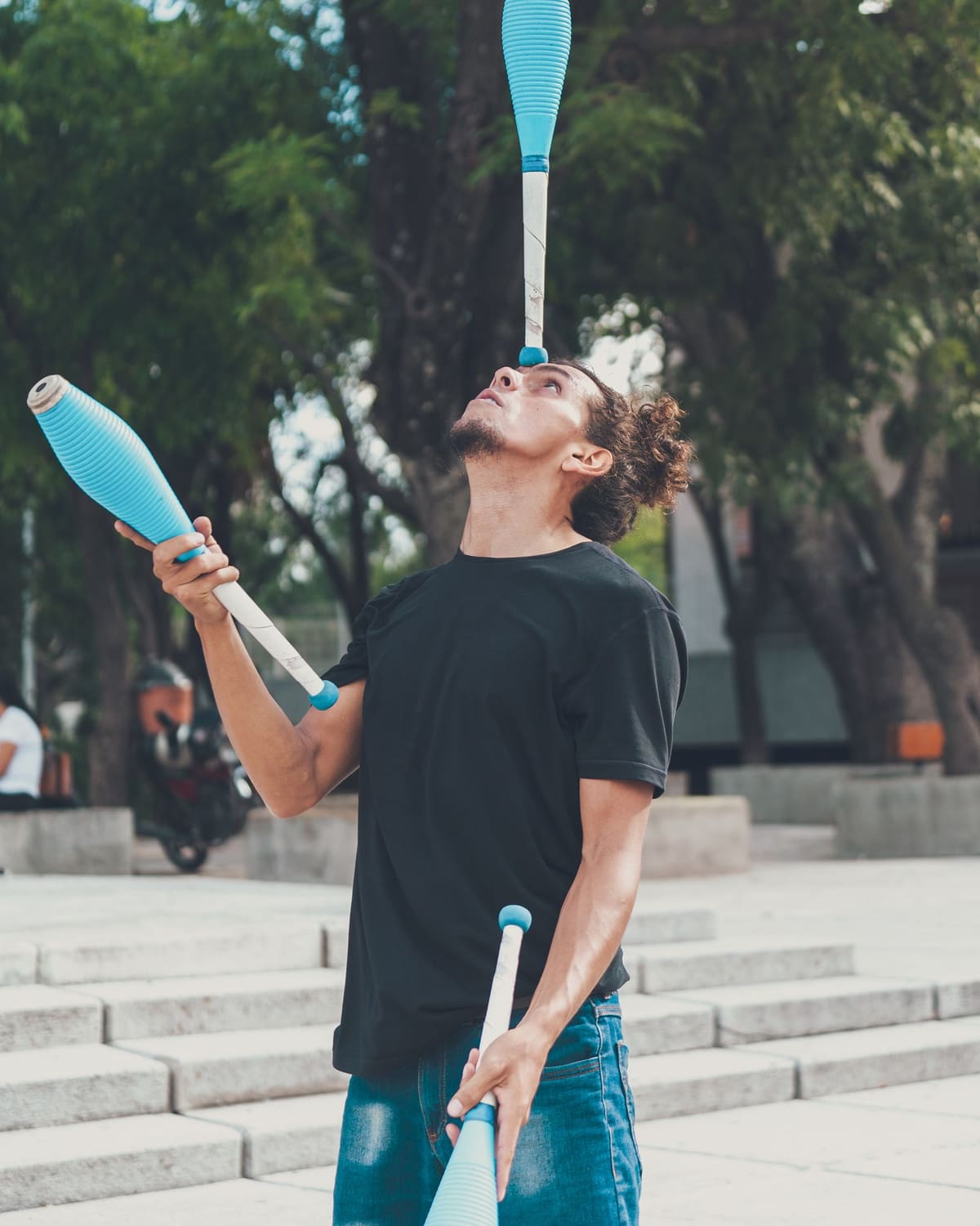 A young man in a black T-shirt and jeans balances a blue-and-white juggling club on his forehead while holding two more, standing outdoors before green trees.