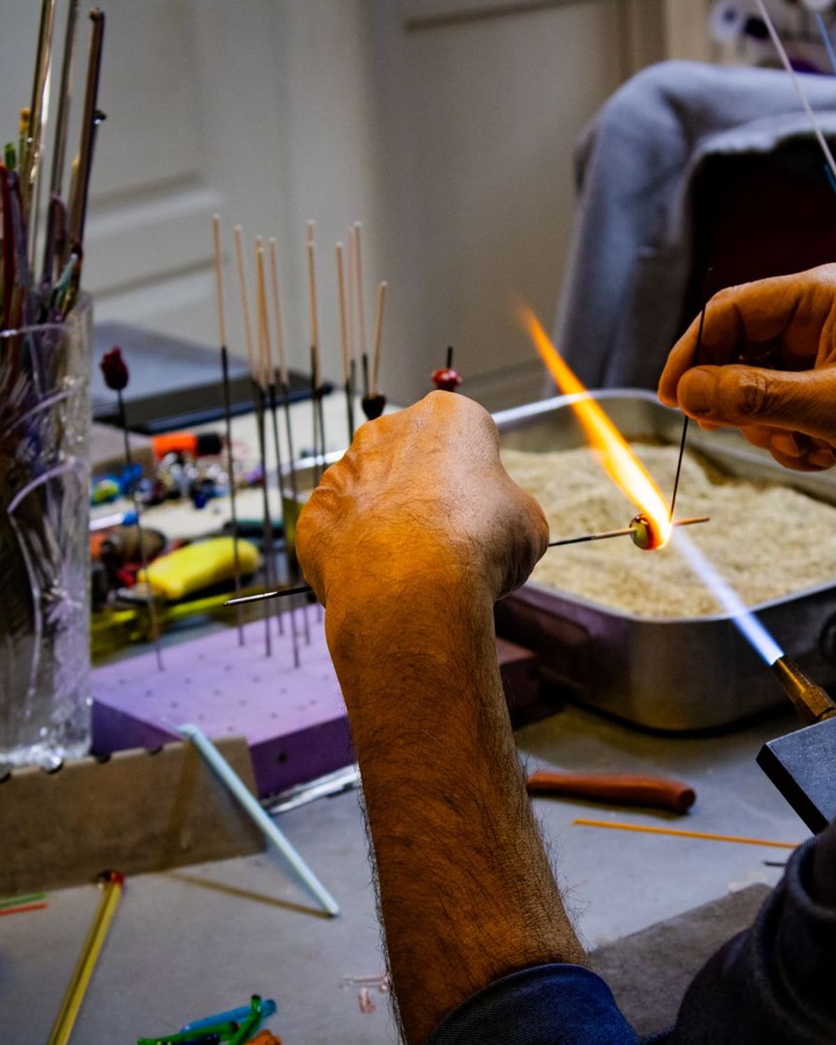 A lampworker's hands turning a glass rod in an orange torch flame, colorful glass rods waiting on the bench