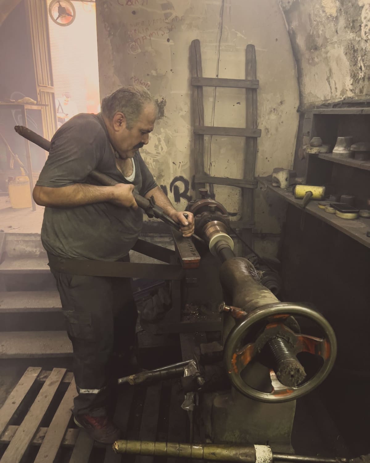 A machinist leaning into a lathe in a warm, worn workshop, working a long metal shaft by hand.