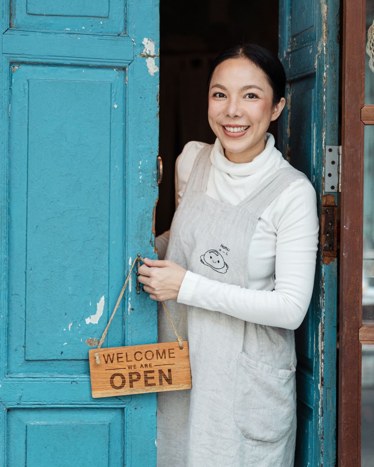 A smiling host in an apron welcoming guests at an open doorway, hanging a wooden open sign in natural light.