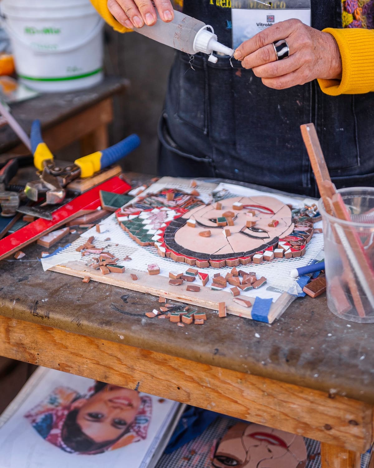 A mosaic maker in an apron setting small colored tiles into a design on a workbench