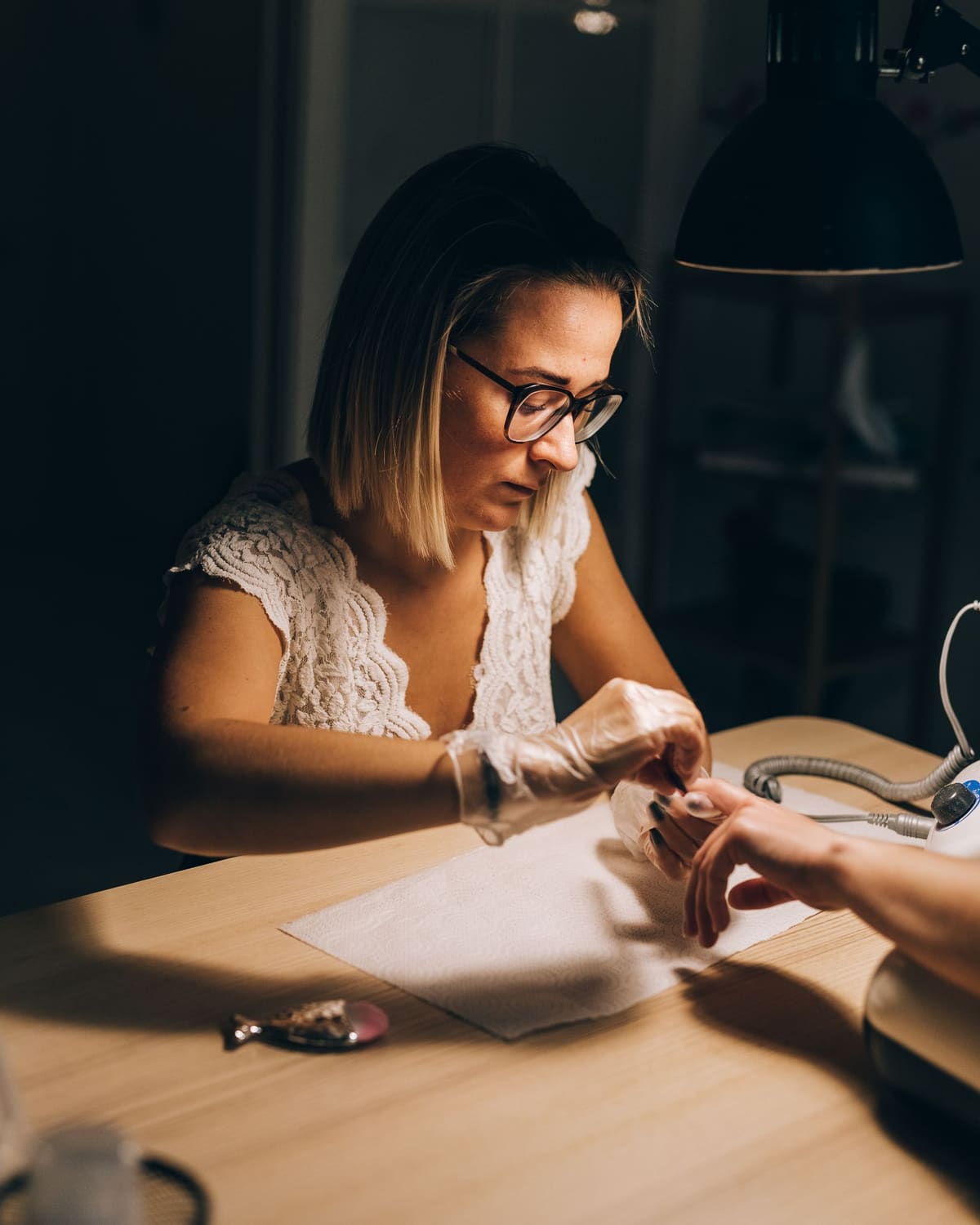 A nail technician under a warm work lamp, gloved hands tending a client's fingers.