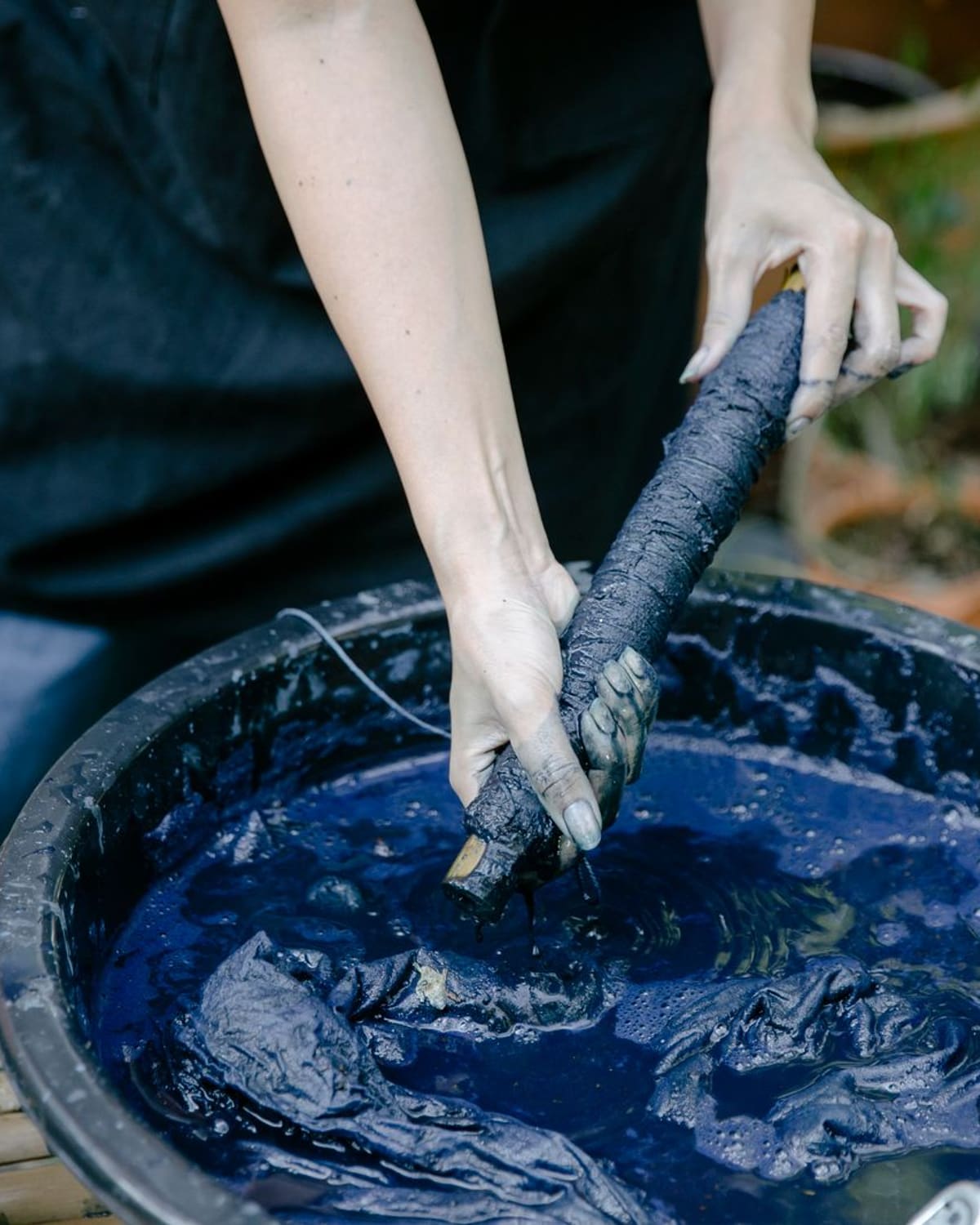 A person's hands dipping a bundled cloth into a vat of deep-blue indigo dye outdoors, fingers stained blue.