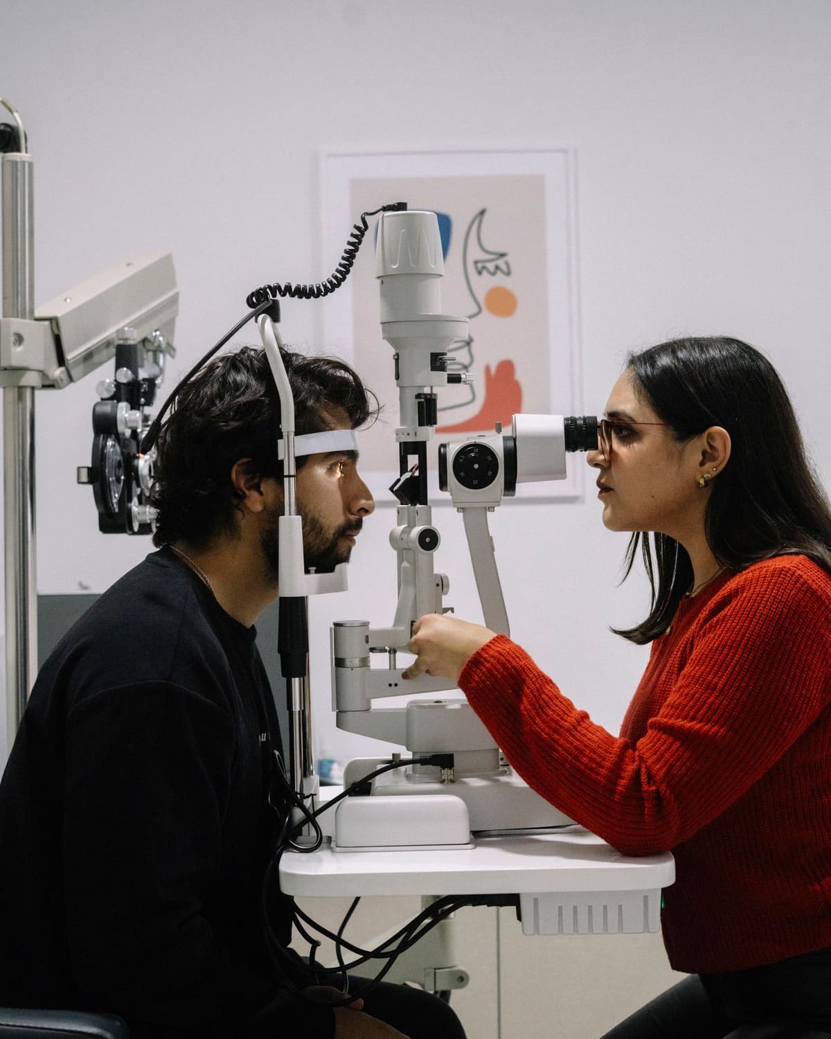 An optometrist at a slit lamp examining a seated patient's illuminated eye, both faces close and focused.