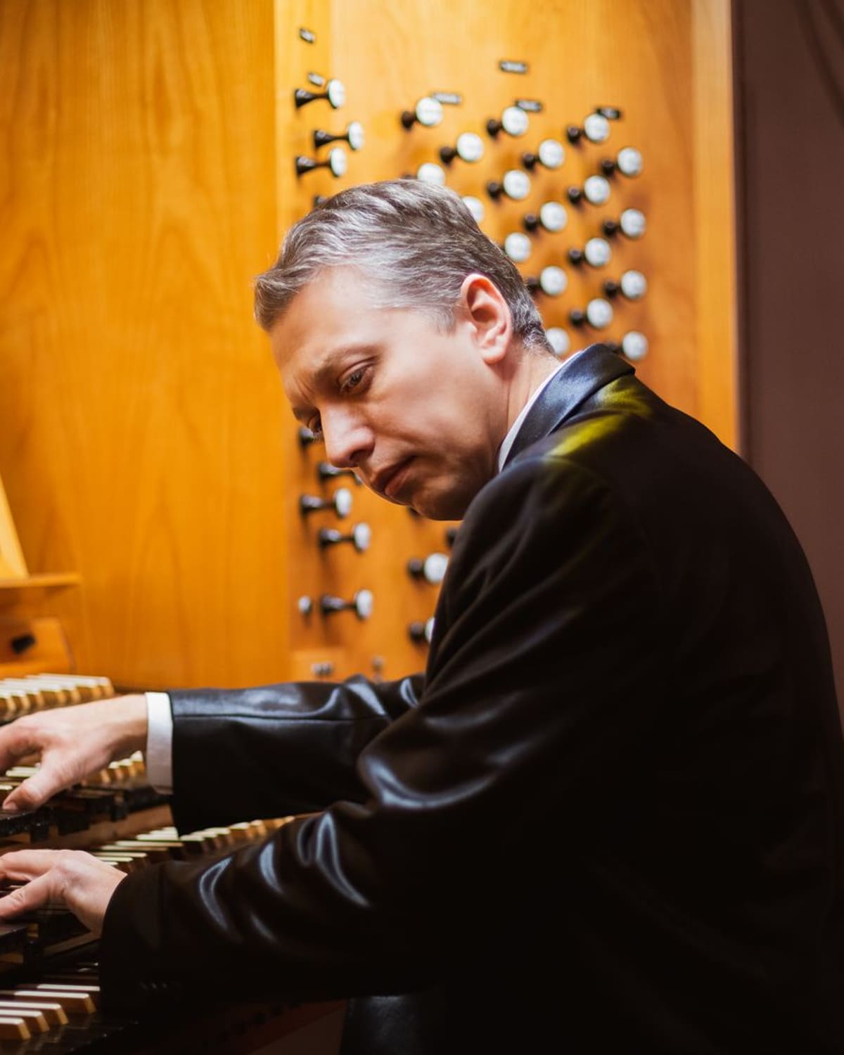 An organist at a pipe-organ console reaches across the stops, warm wood glowing around him.