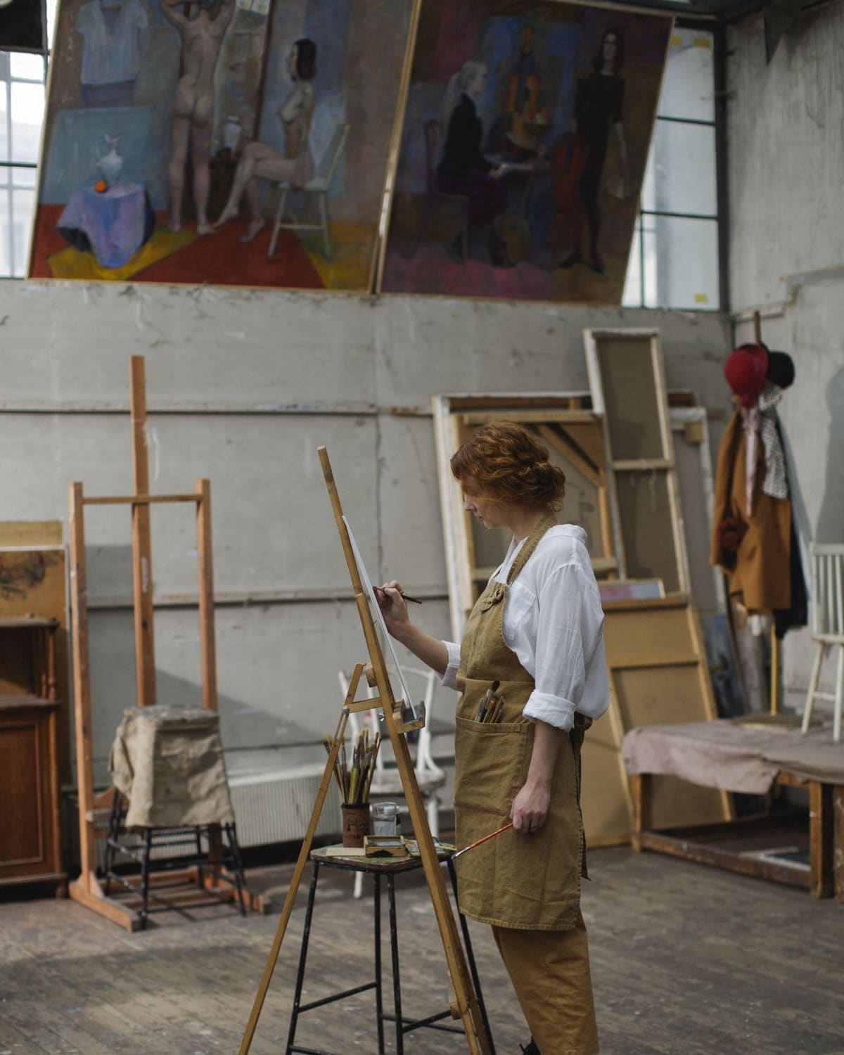 A woman in an apron paints on a canvas at a wooden easel in a bright studio, large paintings on the wall behind her.