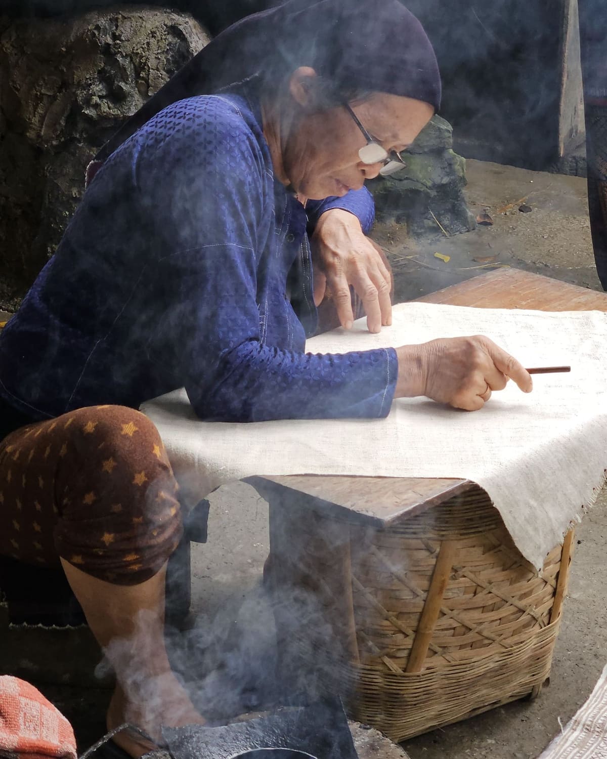 An elderly woman leans over a table smoothing a sheet of handmade paper near a steaming stove.