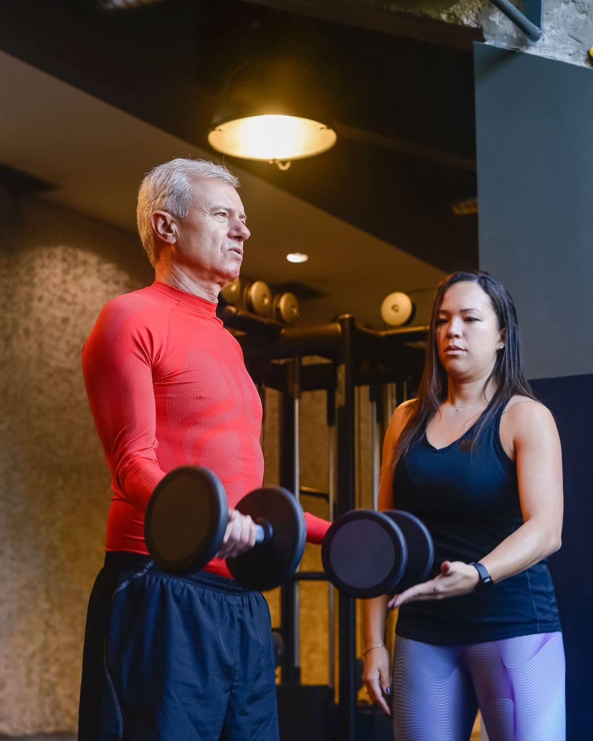 Personal trainer coaching an older man through a dumbbell exercise in a gym.