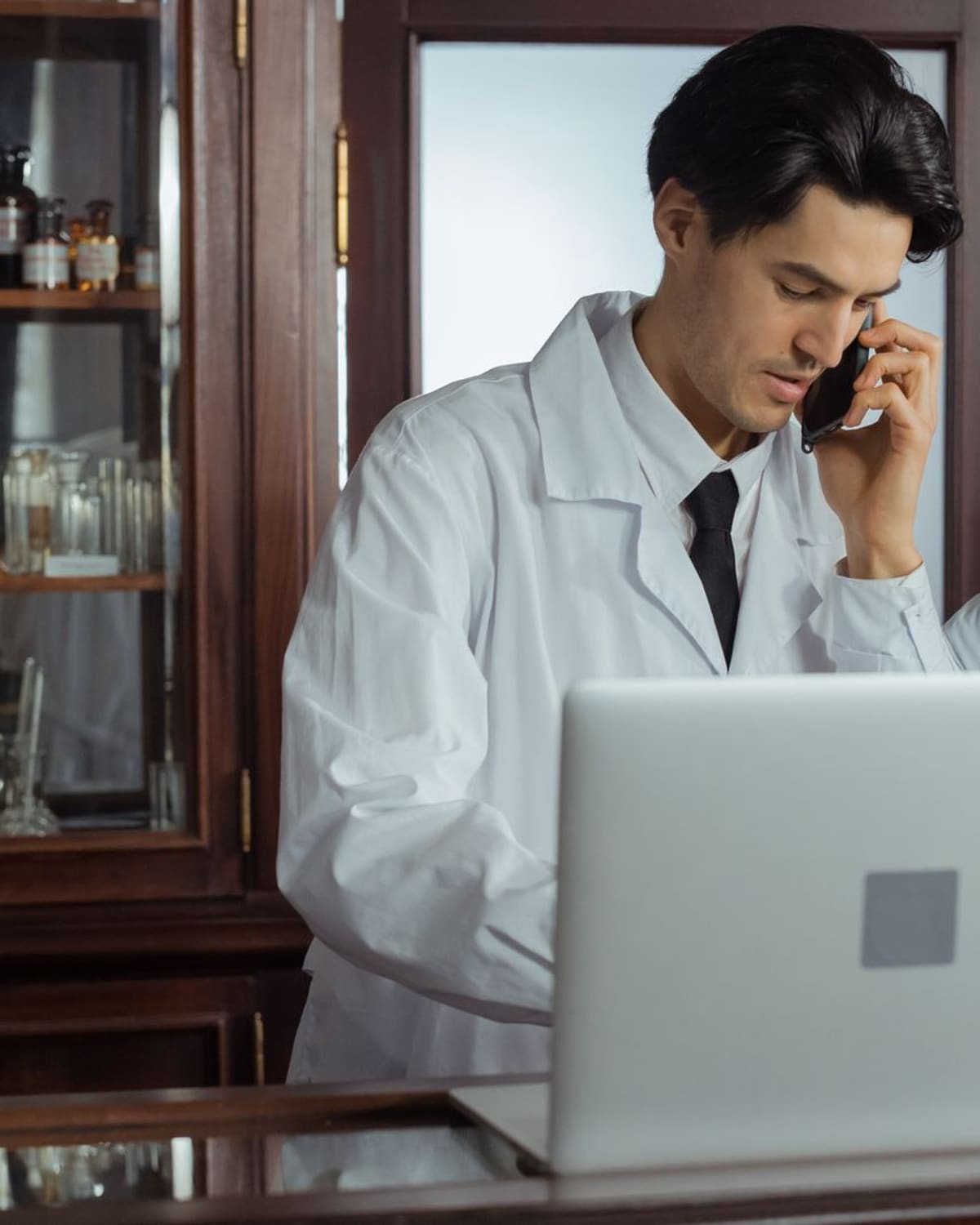 Two pharmacists in white coats working together at a wooden pharmacy counter, one on the phone