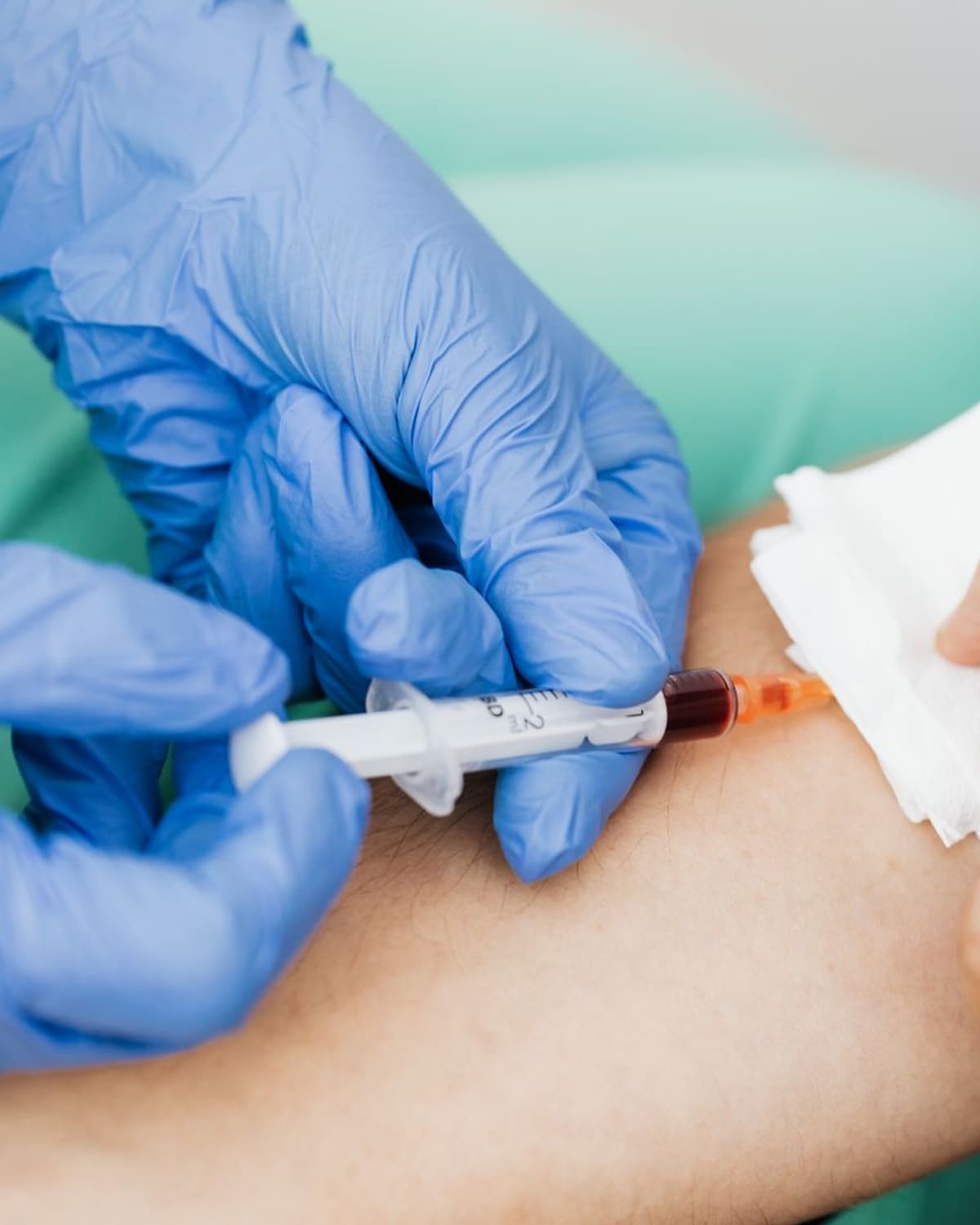 Gloved hands drawing blood from a patients arm into a vial with a small syringe.