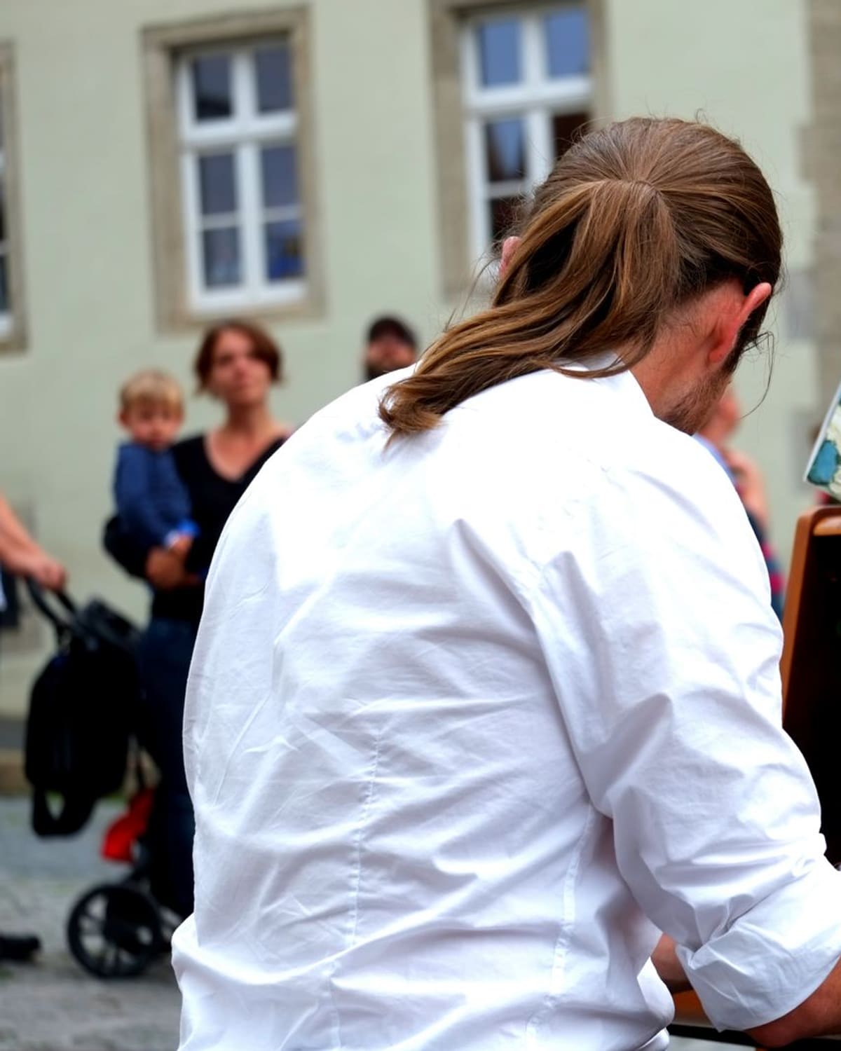A long-haired person in a white shirt plays an open upright piano on a cobblestone street, with people watching in the background under daylight.